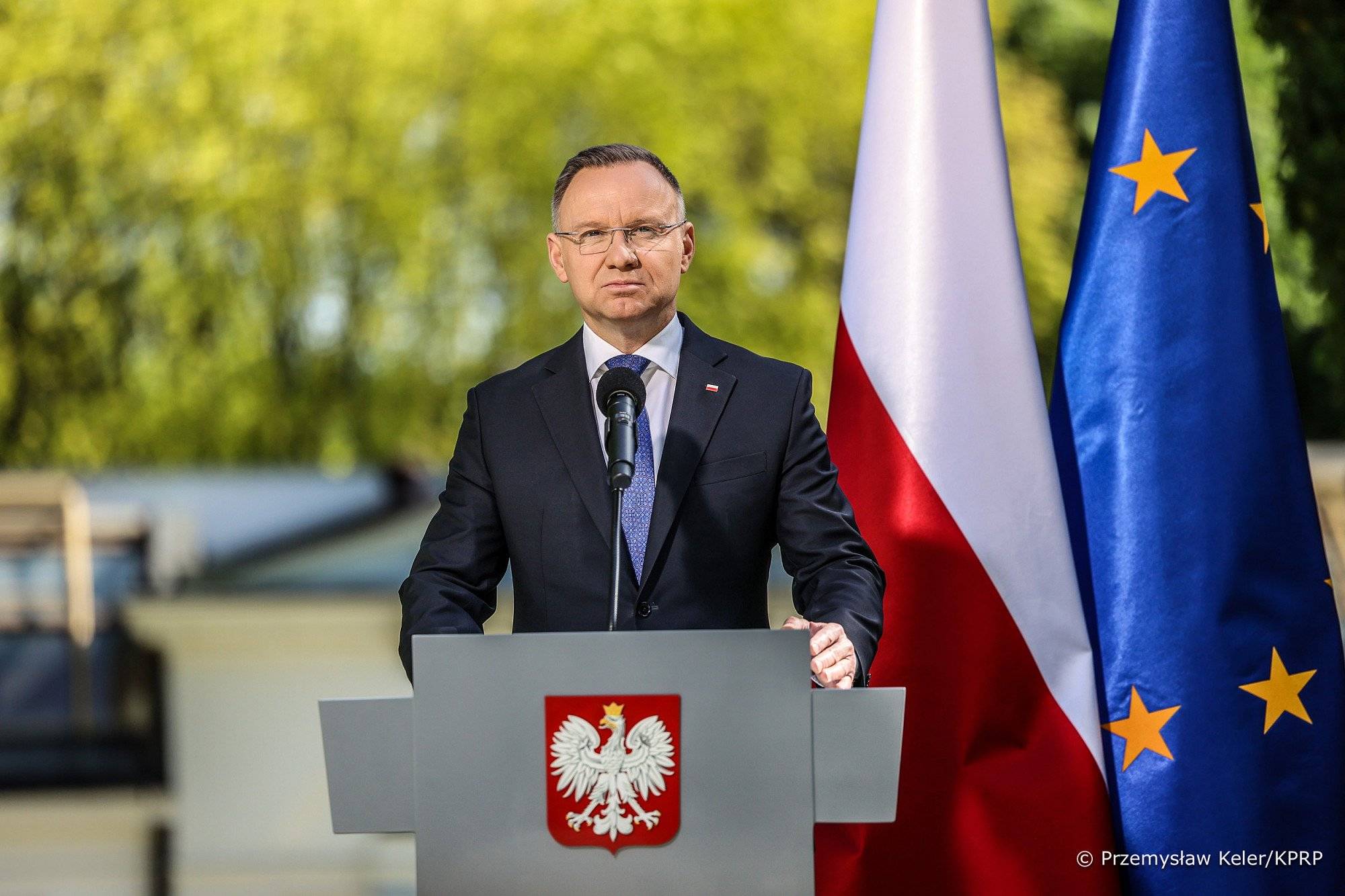 Mężczyzna w czarnym garniturze z niebieskim krawatem stoi przy szarej mównicy z godłem Polski, w tle ogród i zielone drzewa, a po jego lewej stronie flaga Polski i flaga UE.