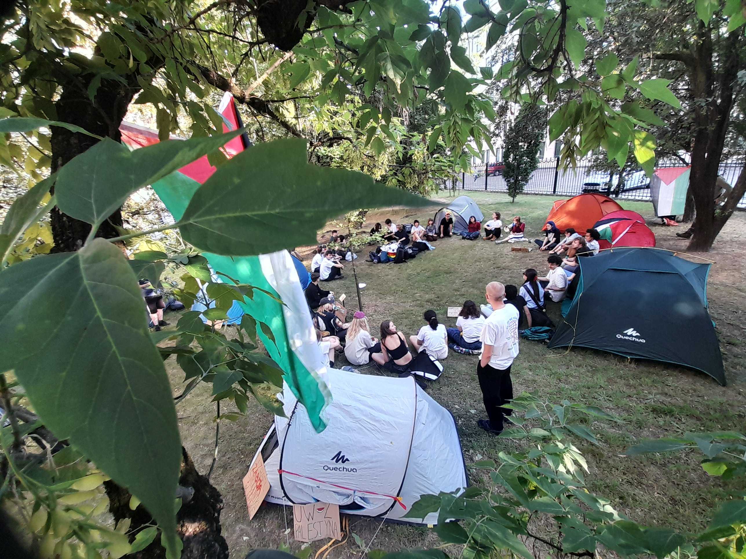 Grupa studentów siedzi na trawie w kółku. Wokół nich namioty, w których będą spać podczas okupacji terenu Uniwersytetu Warszawskiego