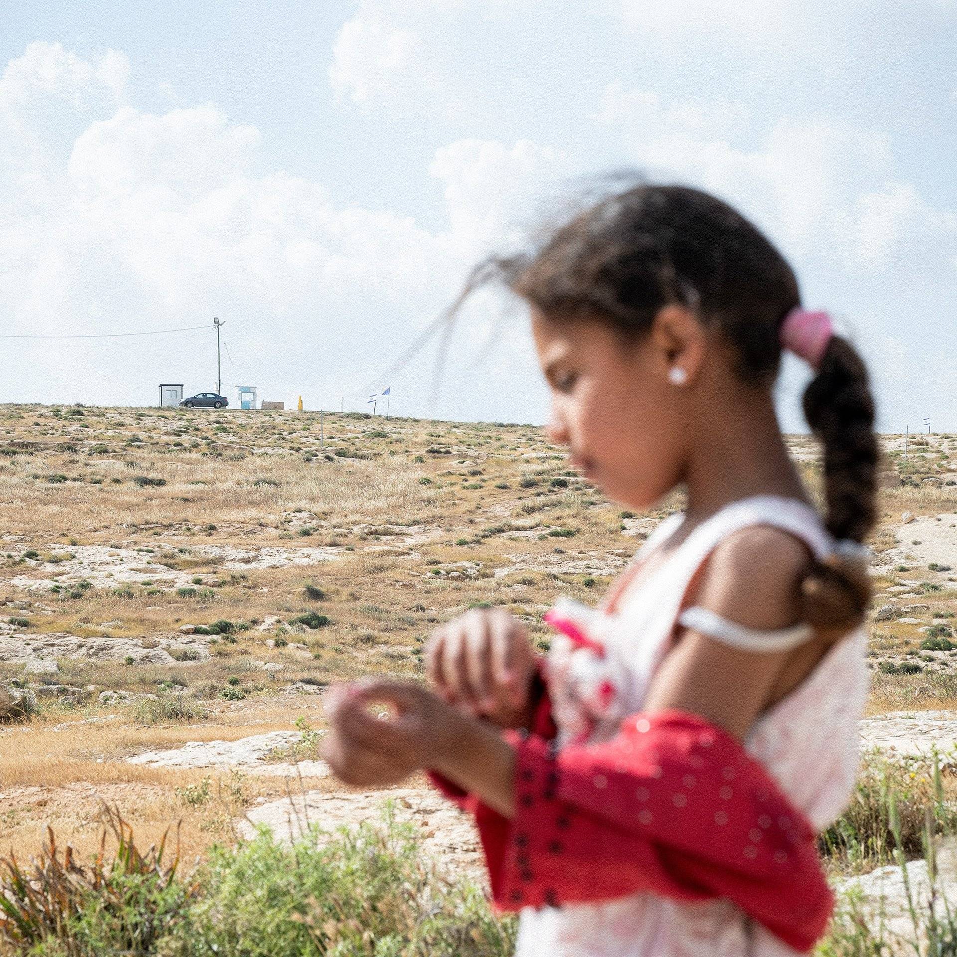 Na pierwszym planie widoczna rozmazana postać palestyńskiej dziewczynki, na drugim planie widoczny wyostrzony posterunek z izraelskimi flagami. Zachodni Brzeg