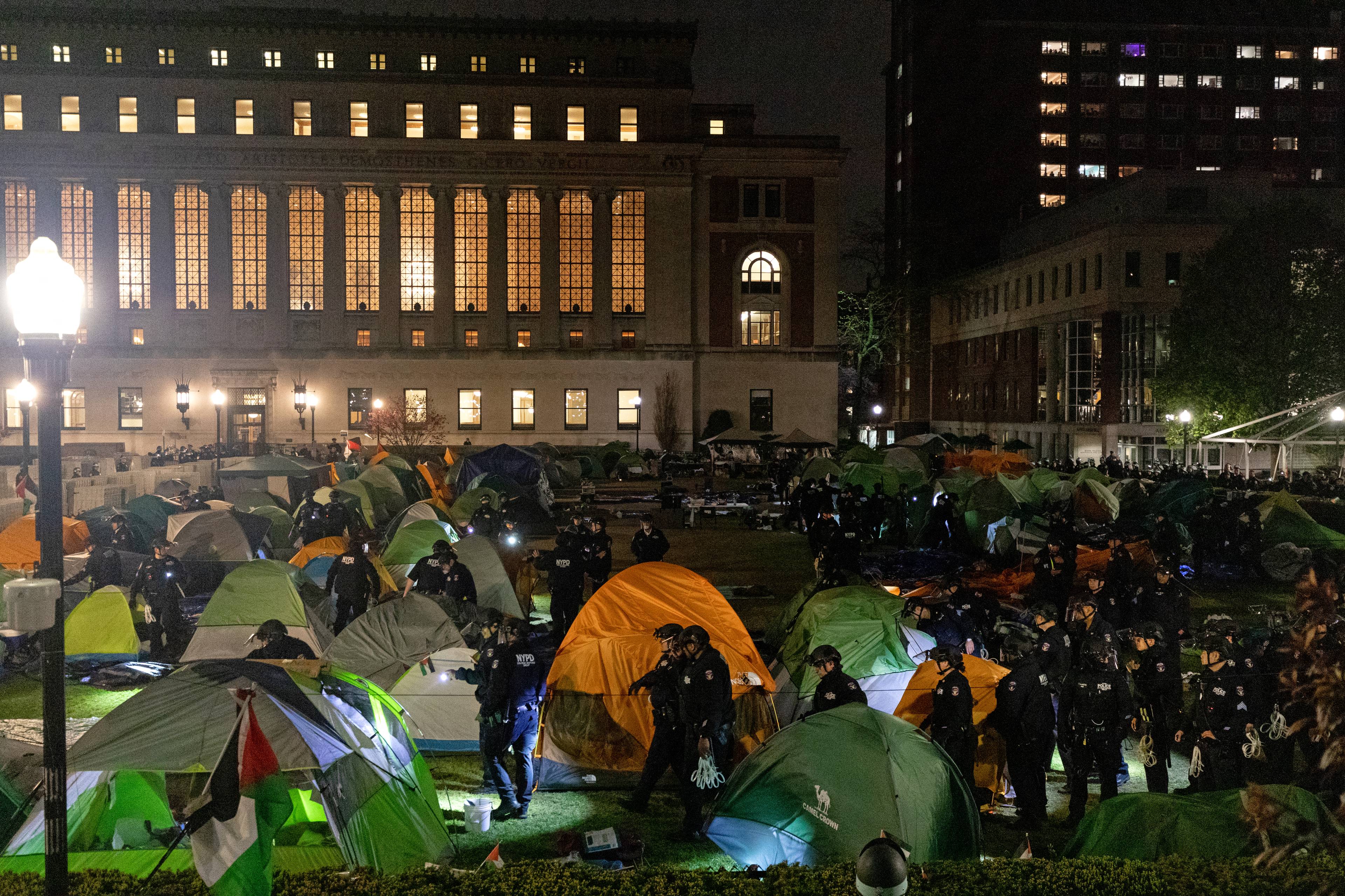 Duży trawnik pomiędzy budynkami Uniwersytetu COlumbia w Nowym Jorku, cały zastawiony namiotami, pomiędzy namiotami widać kordon policji, która zmierza w stronę okupowanego budynku. Jest późny wieczór, ciemno.