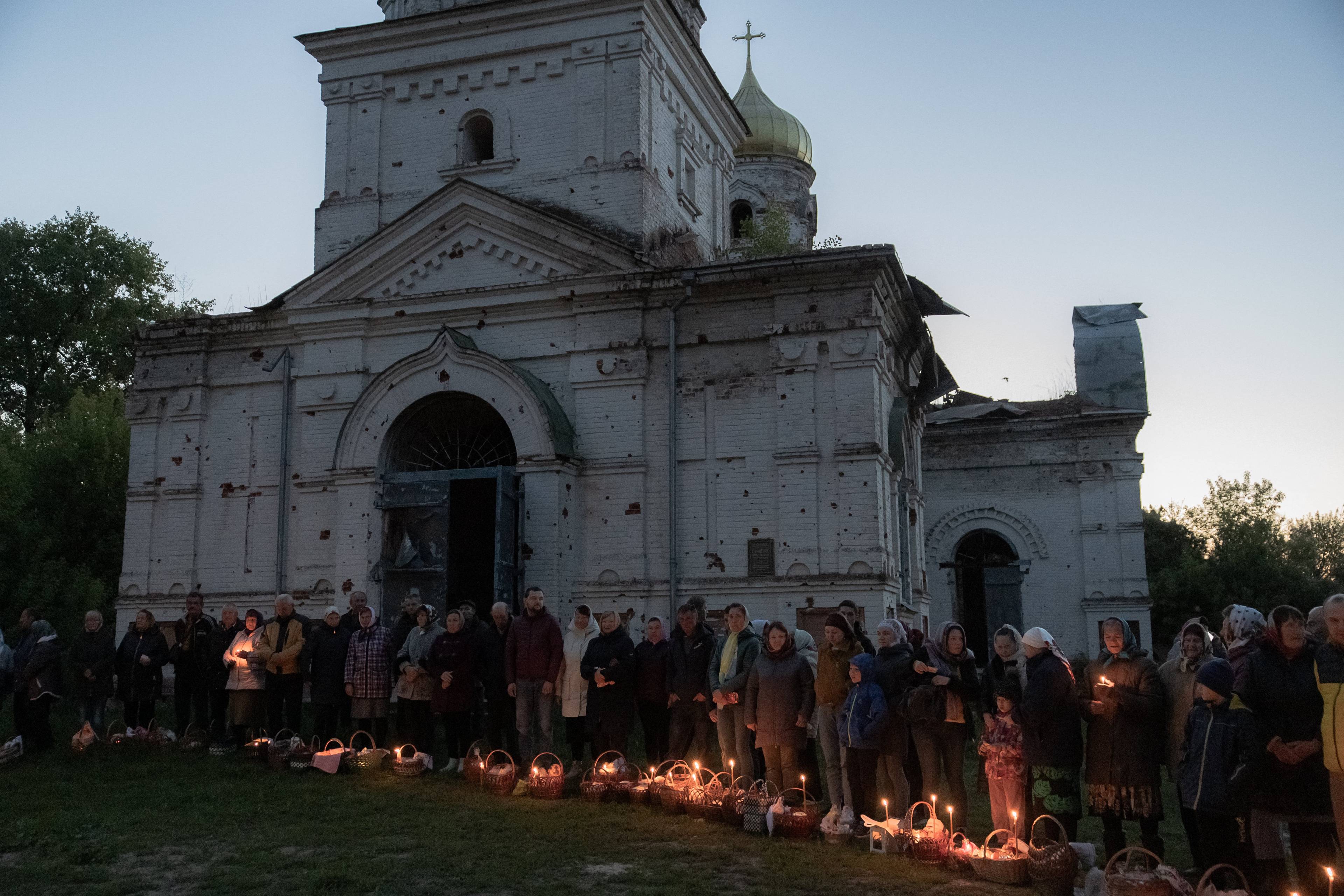 Świt. Na zdjęciu są ludzie, święcą pokarmy obok zniszczonej cerkwi. W koszyczkach palą się świeczki. Wielkanoc prawosławna