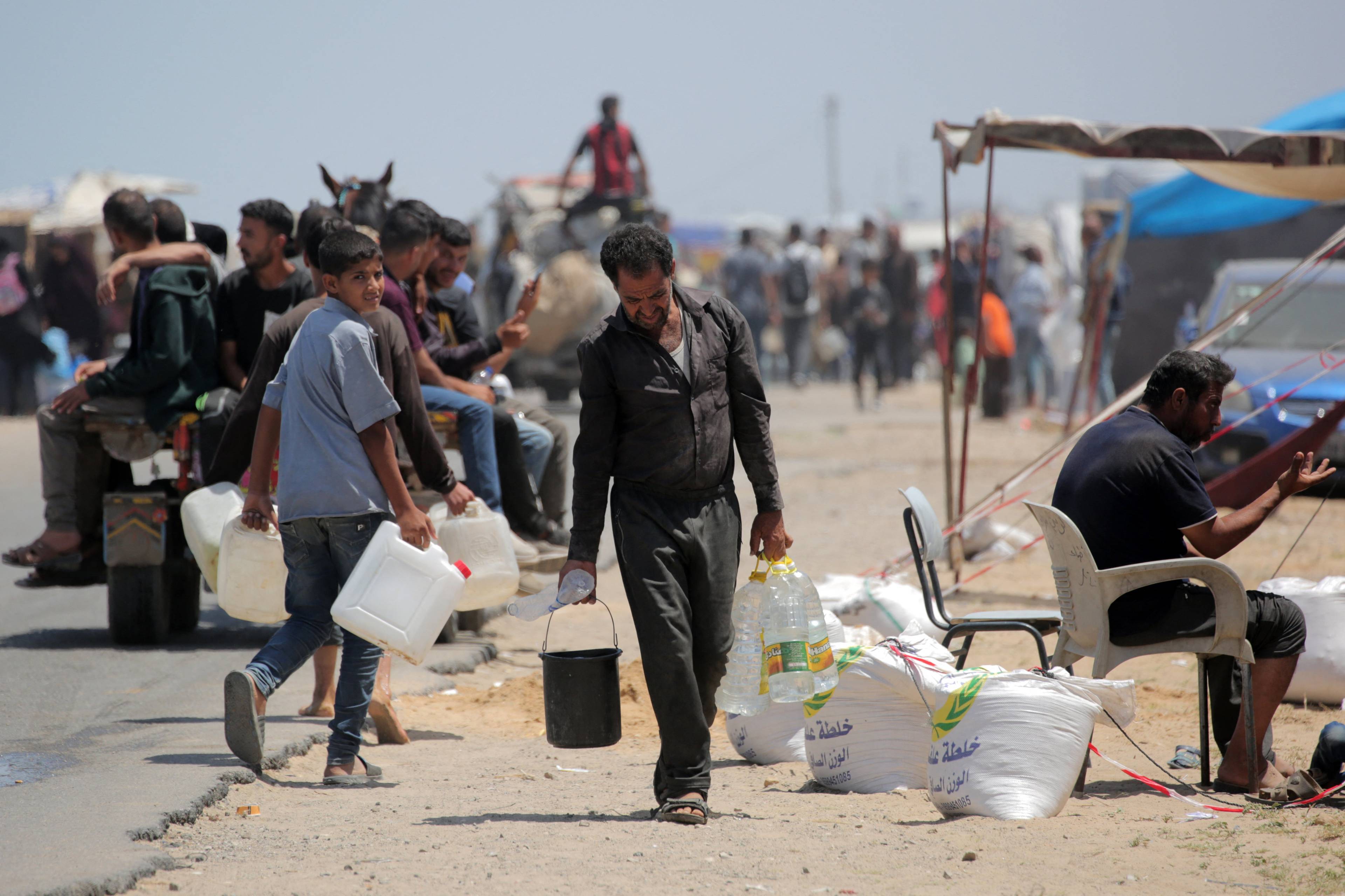 Mężczyzna w Rafah niesie wiadro, prawdopodobnie z wodą. W tle dziesiątki osób w palącym słońcu