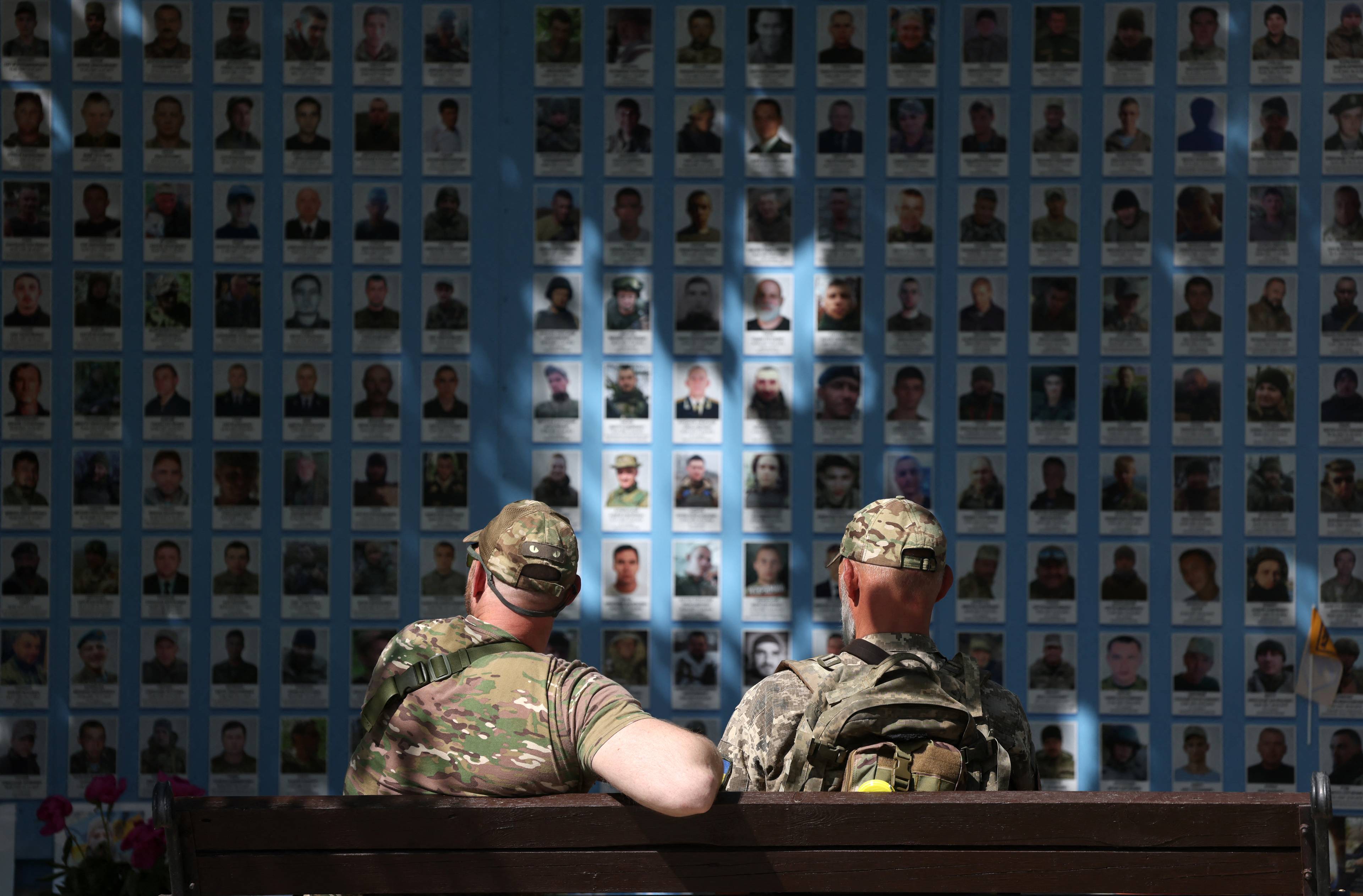 dwóch mężczyzn w mundurach siedzi przed ścianą ze zdjęciami poległych żołnierzy