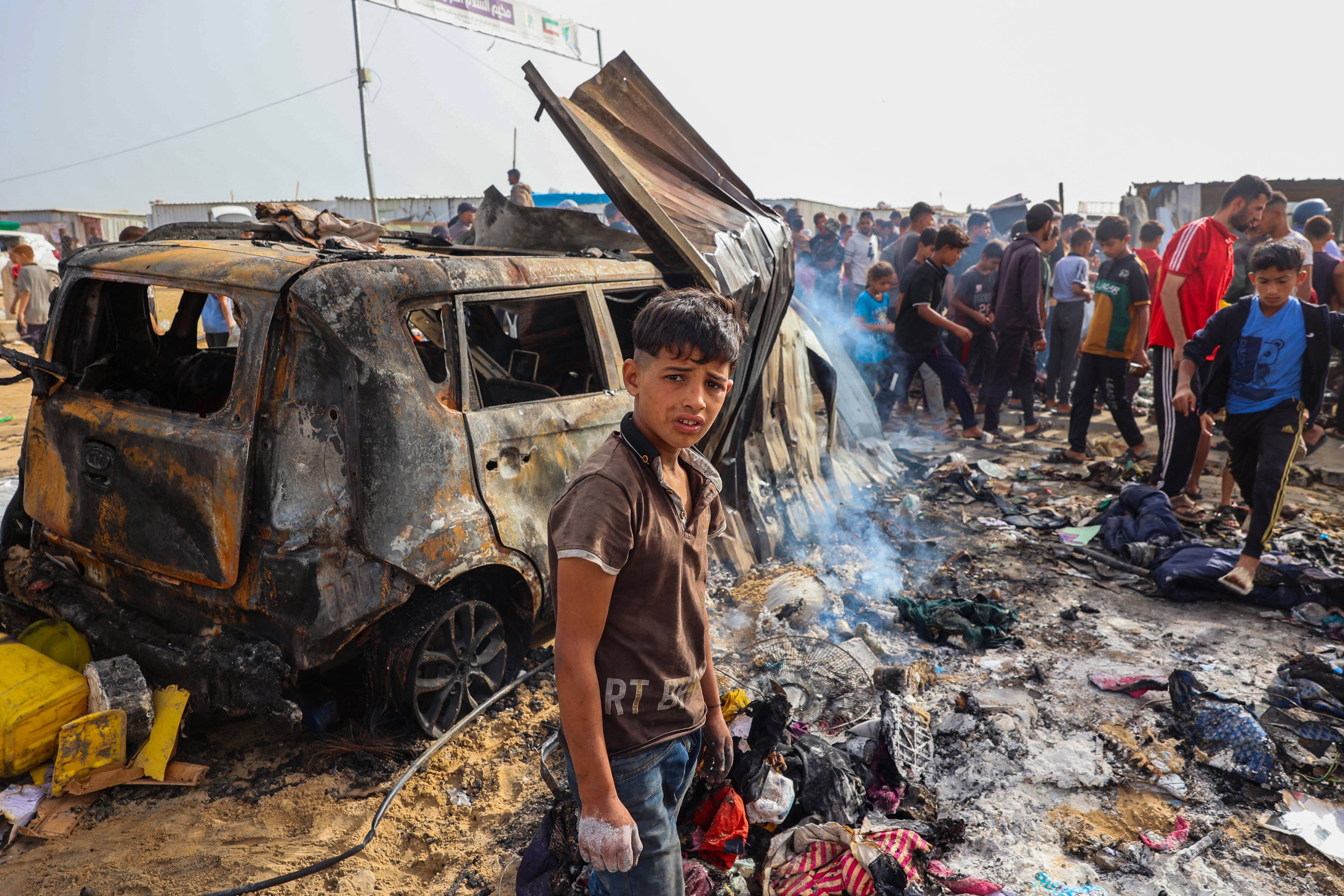 Nastoletni chłopiec stoi na tle spalonego samochodu, pośród pozostałości po namiotach spalonych po izraelskim ataku w Rafah