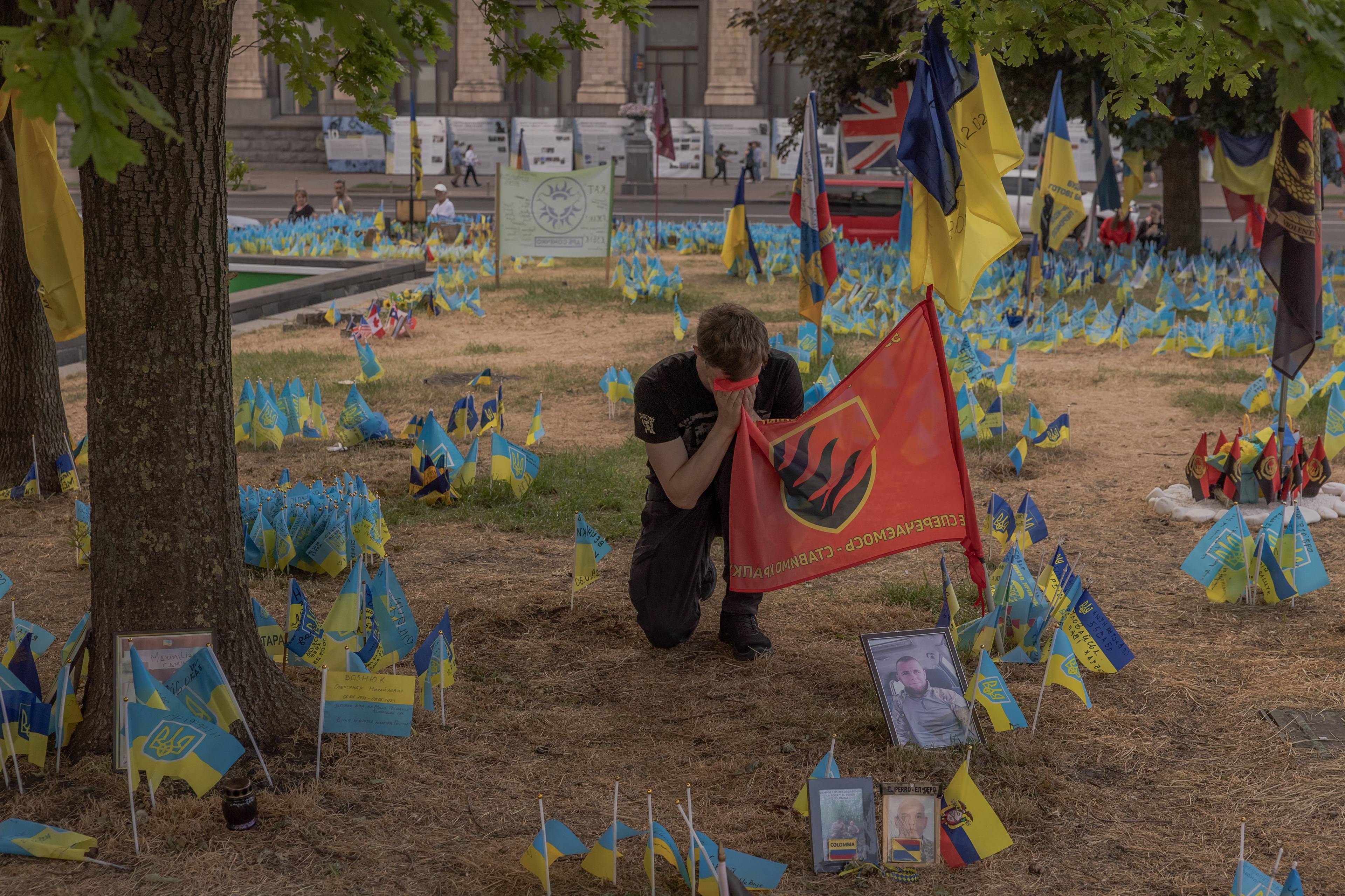 żołnierz klęczy i całuje flagę brygady pośród zatkniętych w ziemię flag ukraińskich