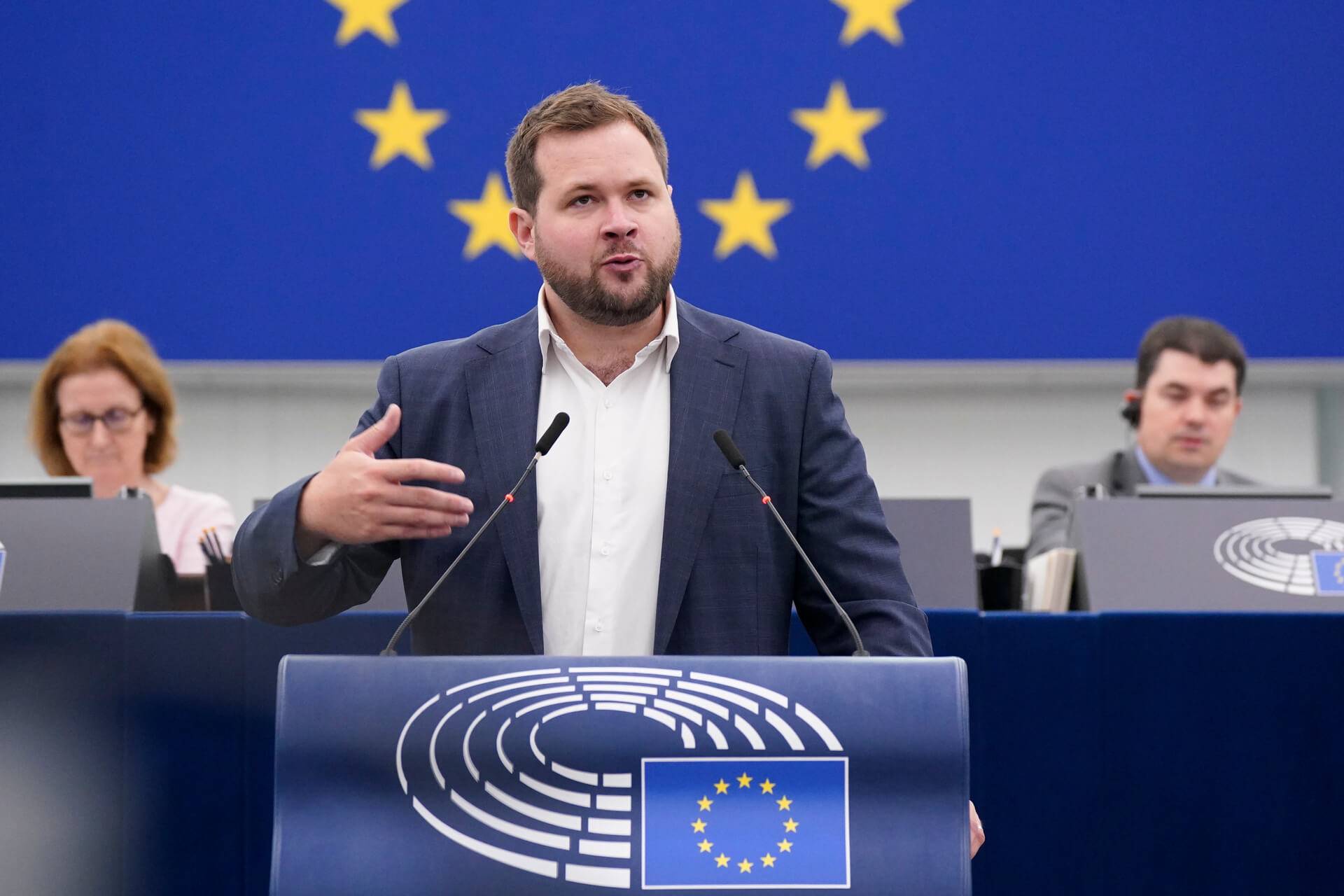 Mężczyzna z krótkimi włosami ciemny blond i krótką brodą stoi za mównicą w sali plenarnej Parlamentu Europejskiego, w tle za nim europejska flaga z gwiazdkami, gestykuluje prawą ręką i przemawia do zebranych europosłów.