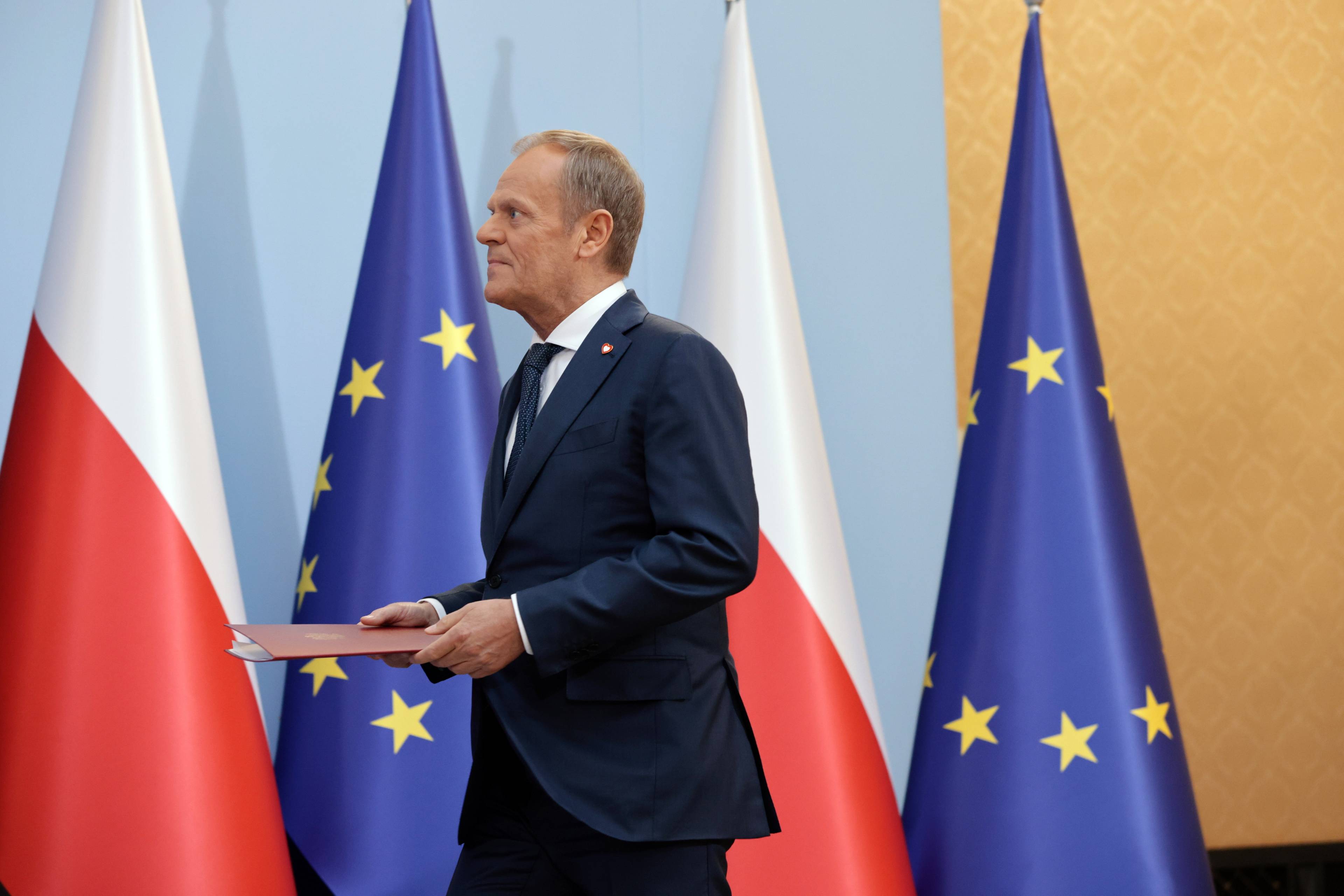 Donald Tusk idzie w lewą stronę w dłoniach trzyma teczkę z dokumentem. W tle flagi Polski oraz Unii Europejskiej
