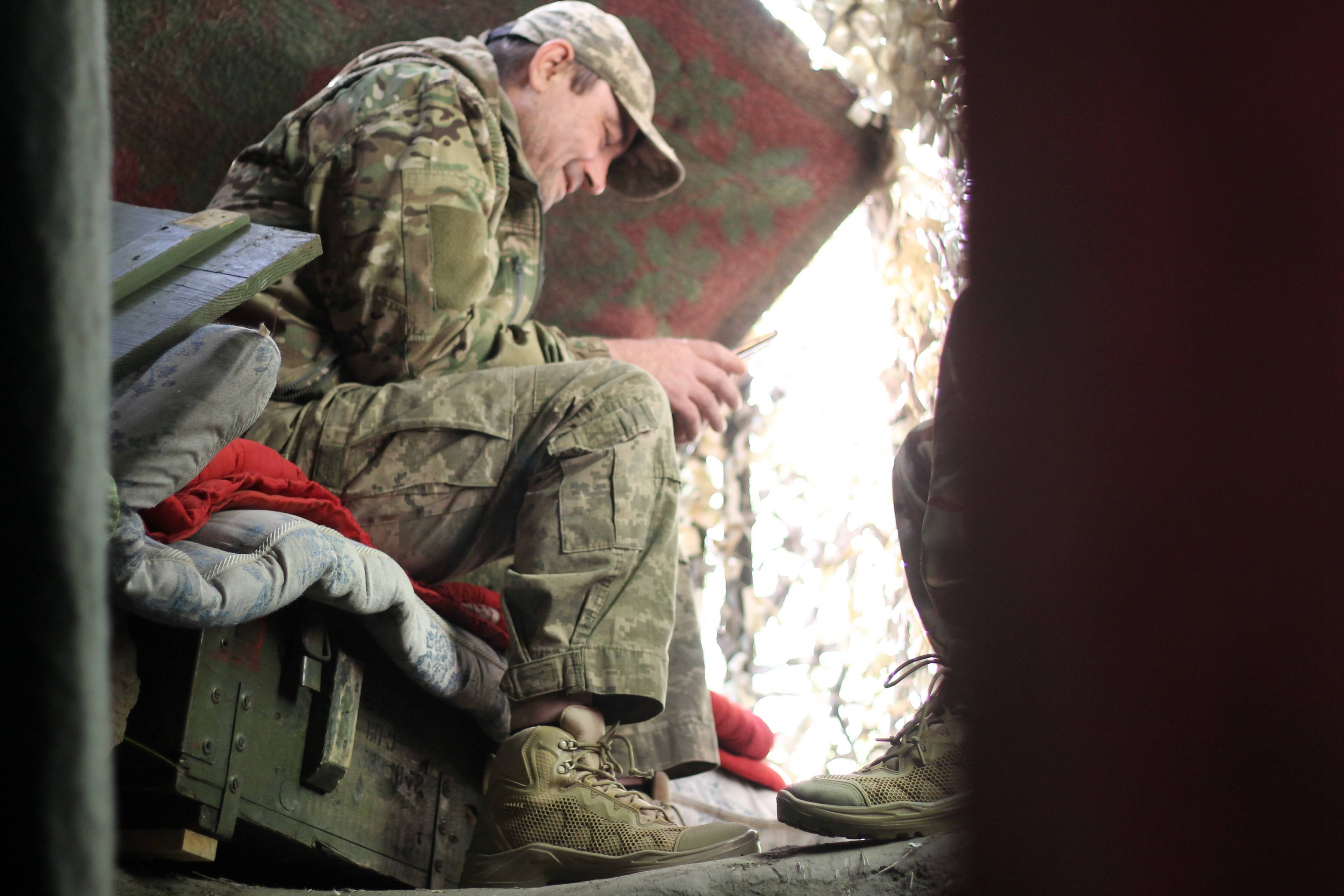 Żołnierz siedzi w ziemiance na drewnianej skrzyni po amunicji i przegląda telefon.