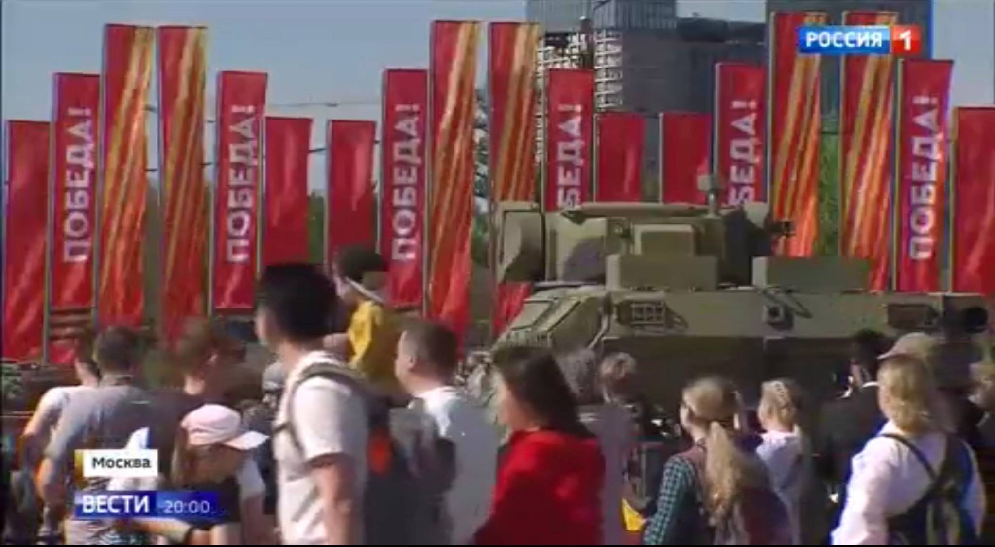 Ludzie oglądają sprzęt wojskowy, w tle - czerwone flagi z rosyjskim napisem 'zwycięstwo"