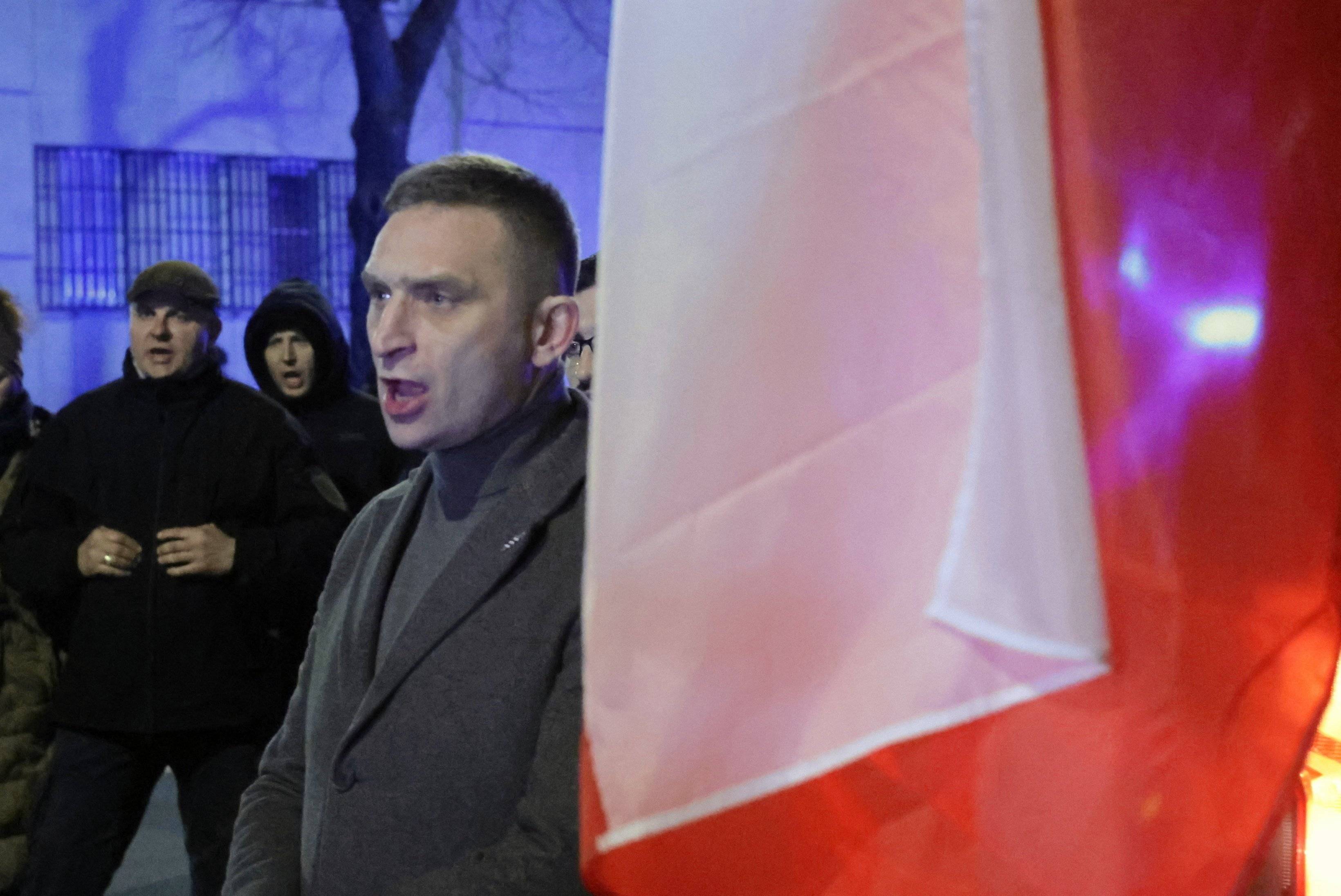 Ubrany w płaszcz mężczyzna krzyczy na ulicy, obok niego polska flaga