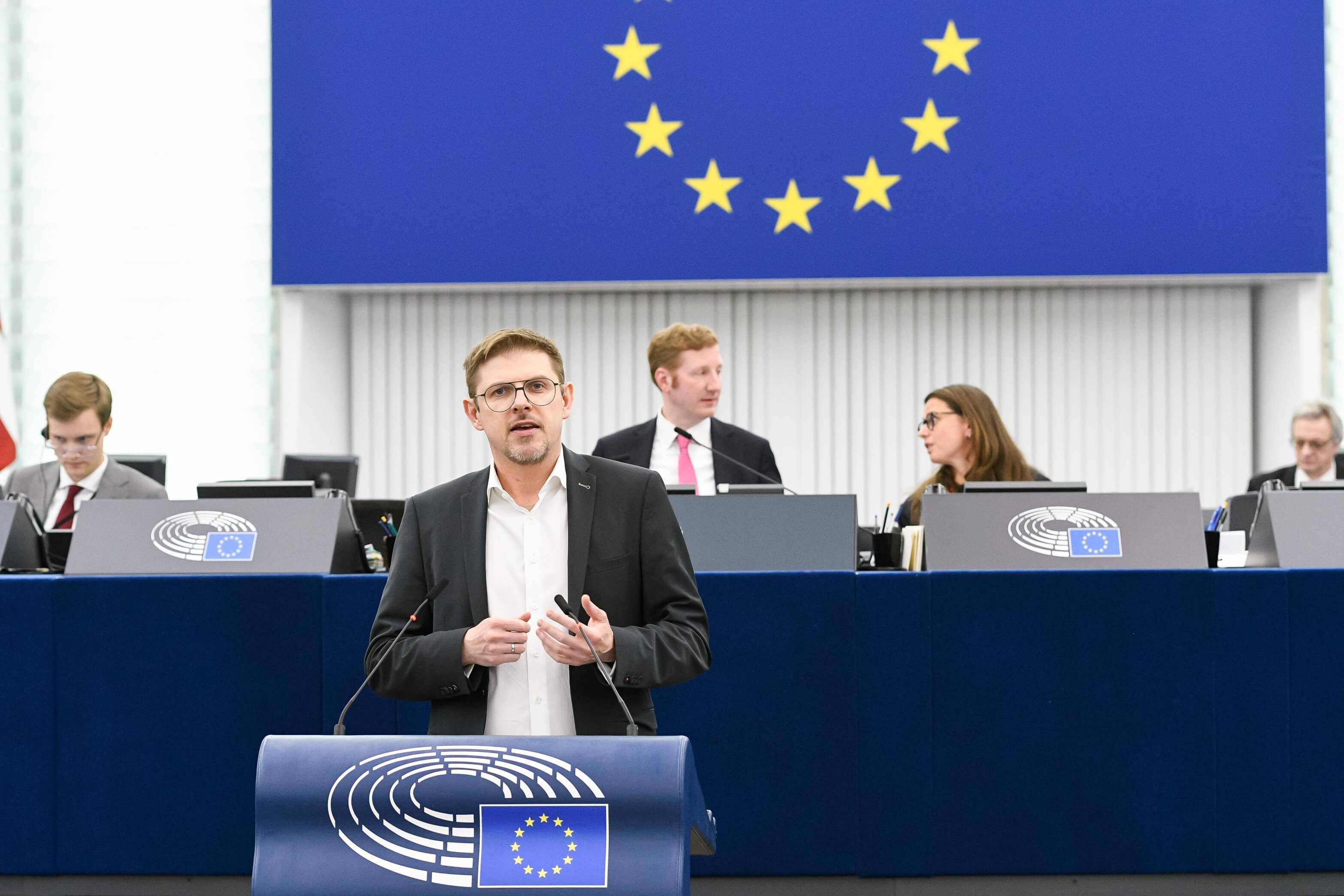 Mężczyzna z krótkimi włosami ciemny blond w okularach, białej koszuli i czarnej, casualowej marynarce stoi przy mównicy w sali plenarnej Parlamentu Europejskiego, w tle widać kawałek unijnej flagi oraz dwie osoby - kobietę i mężczyznę - innych europosłów