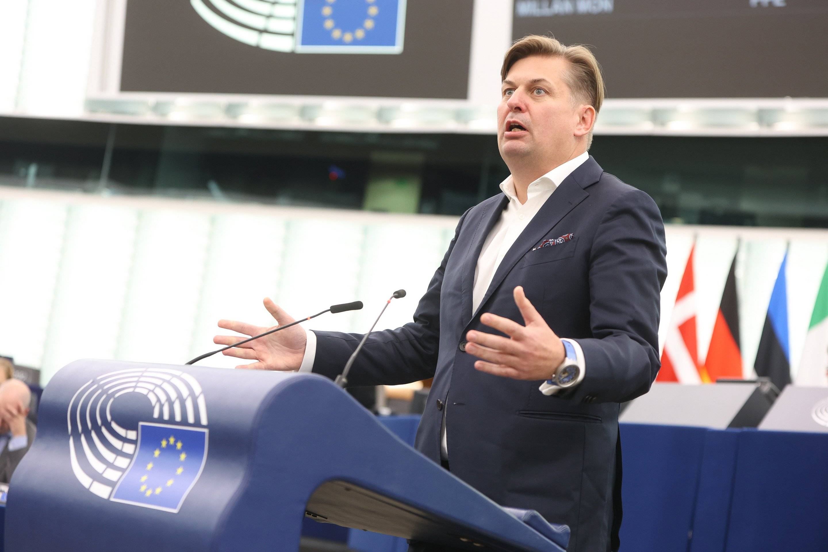 Mężczyzna w czarnej marynarce i białej koszuli bez krawata stoi na podium w sali plenarnej Parlamentu Europejskiego w Brukseli i energicznie gestykuluje. Jego twarz wyraża oburzenie, może zdziwienie