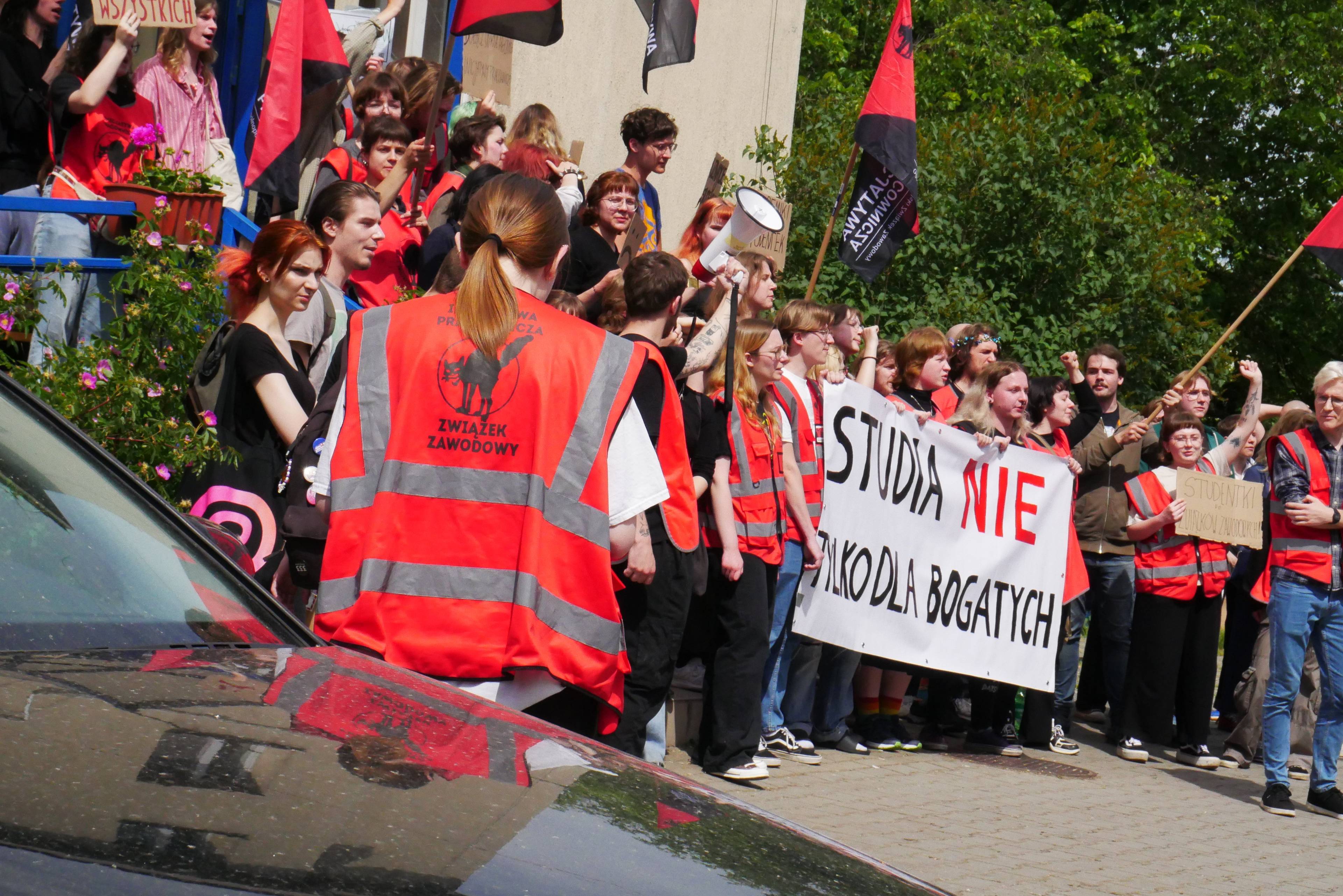 studenci protestujący przed budynkiem akademika w czerwonych kamizelkach. baner z napisem "studia nie tylko dla bogatych"