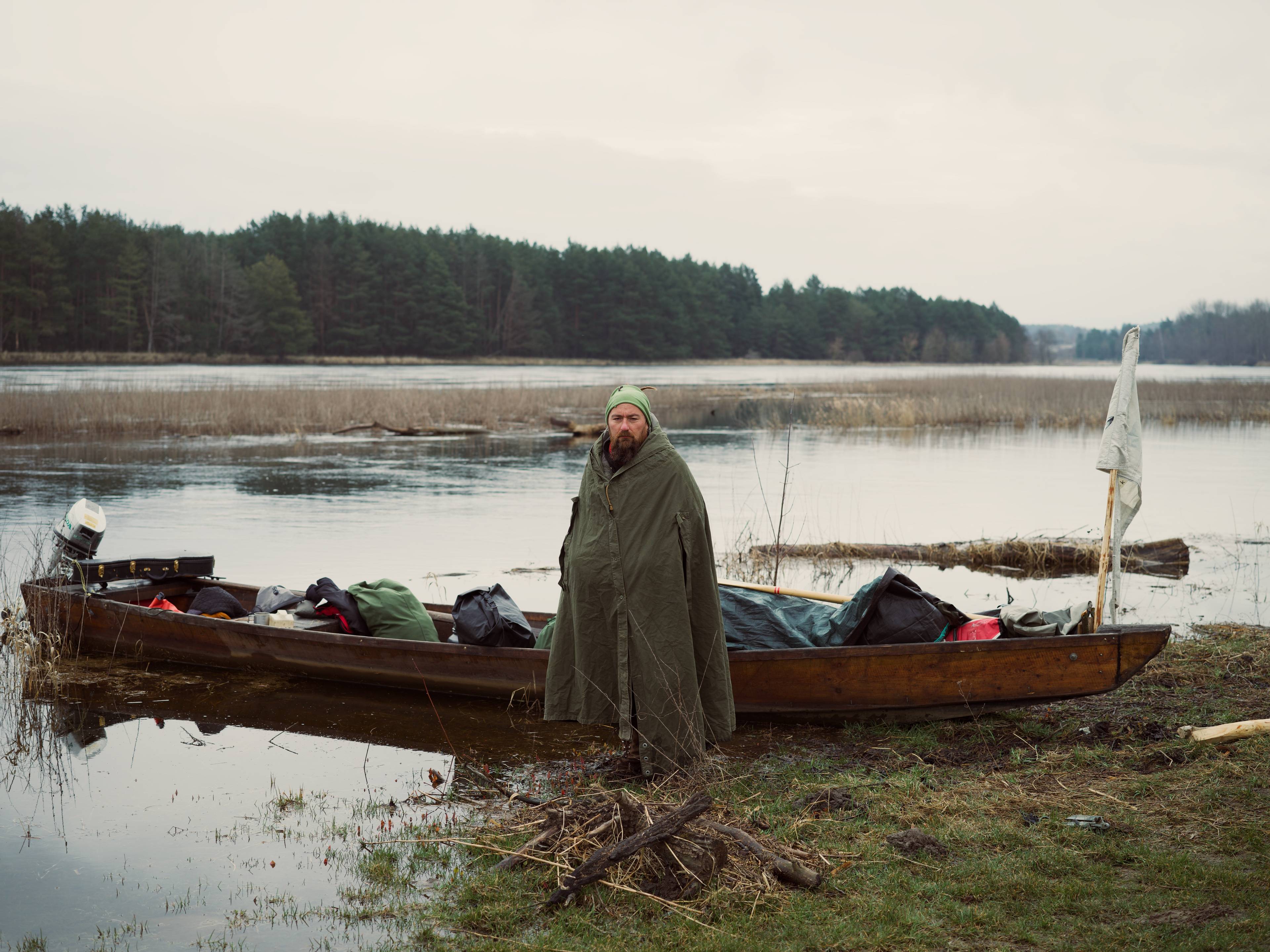 Michał Zygmunt stoi w przeciwdeszczowym płaszczu przy drewnianej łódce pełnej bagaży. Bug - Odra