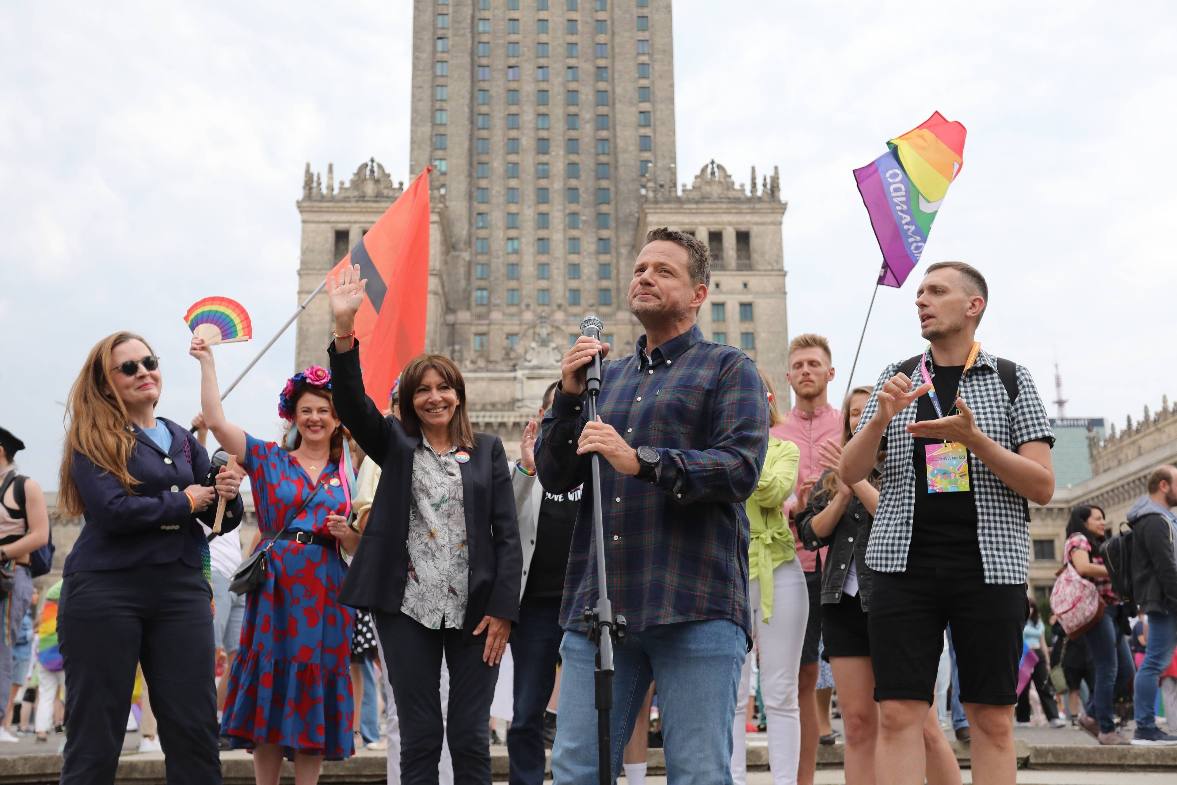 Rafał Trzaskowski w grupie kilku osób, z prawej tęczowa flaga, z tyłu Pałac Kultury i Nauki