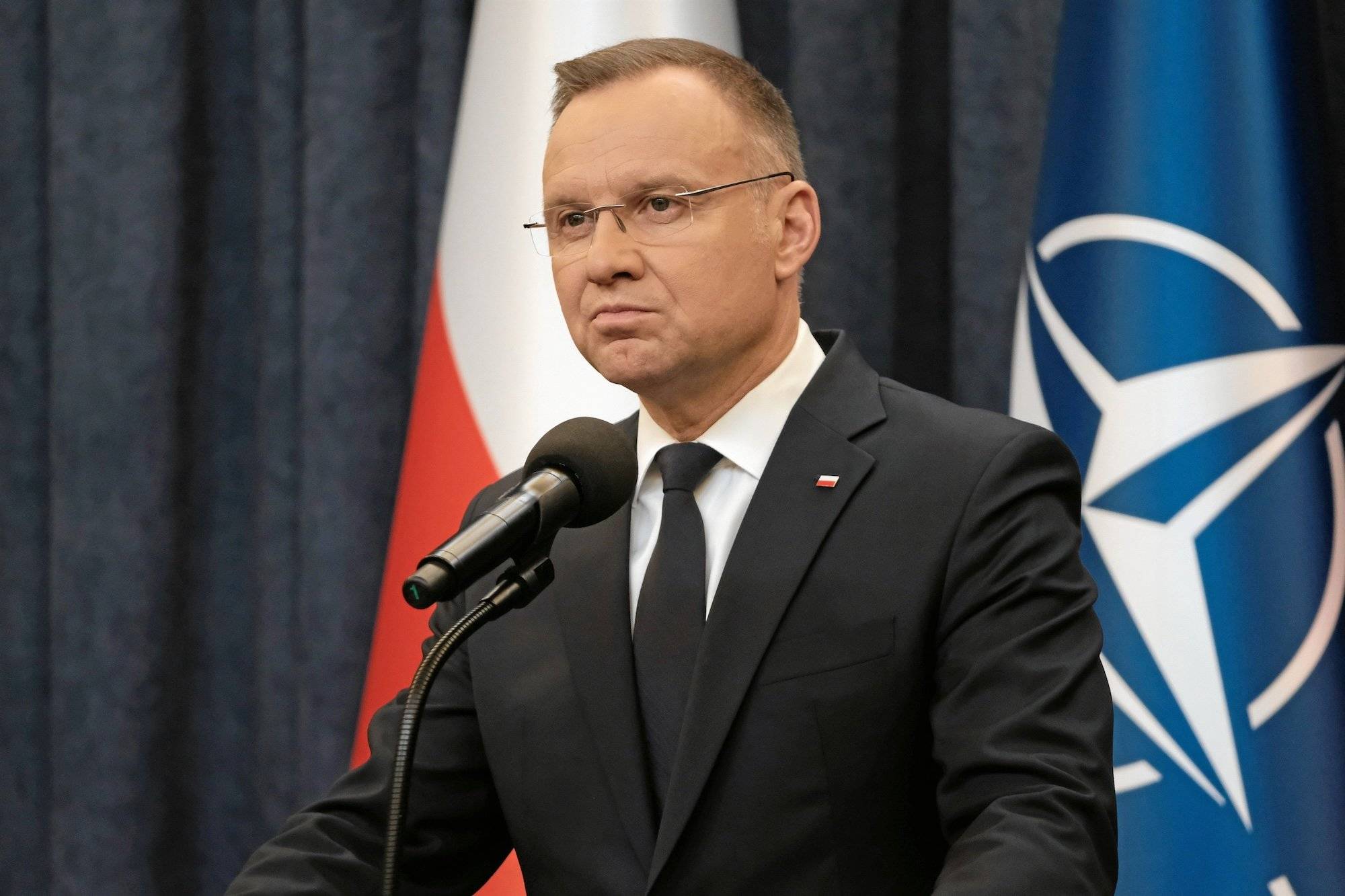 Andrzej Duda w okularach stoi przy mikrofonie na tle flag Polski i NATO