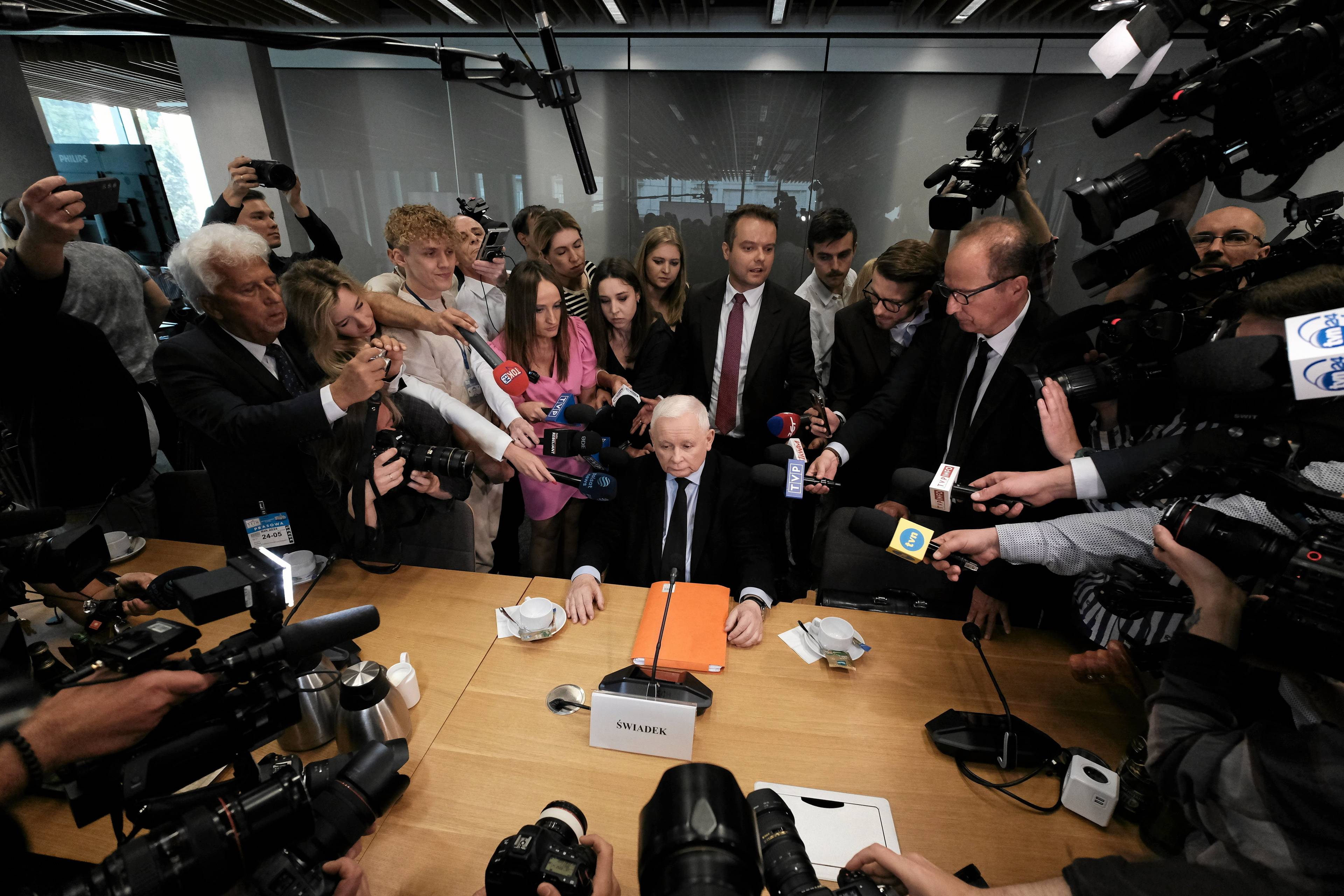 Starszy siwy mężczyzna siedzący przy stole, otoczony dziennikarzami z mikrofonami