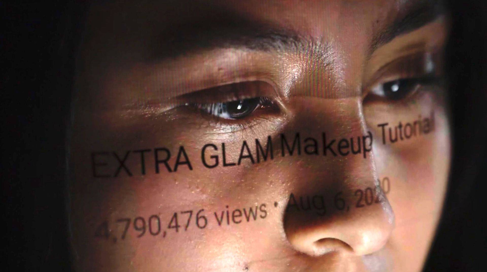 Twarz kobiety, na której odbija się obraz z ekranu komputera z napisem w j. angielskim Extra Glam Make Up Tutorial