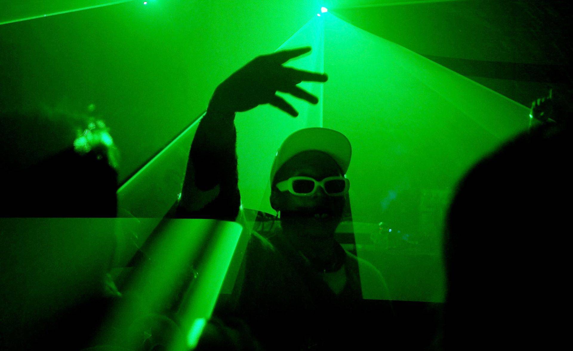 Mężczyzna w białej czapce i okularach przeciwsłonecznych tańczy na tle zielonego światła. Techno