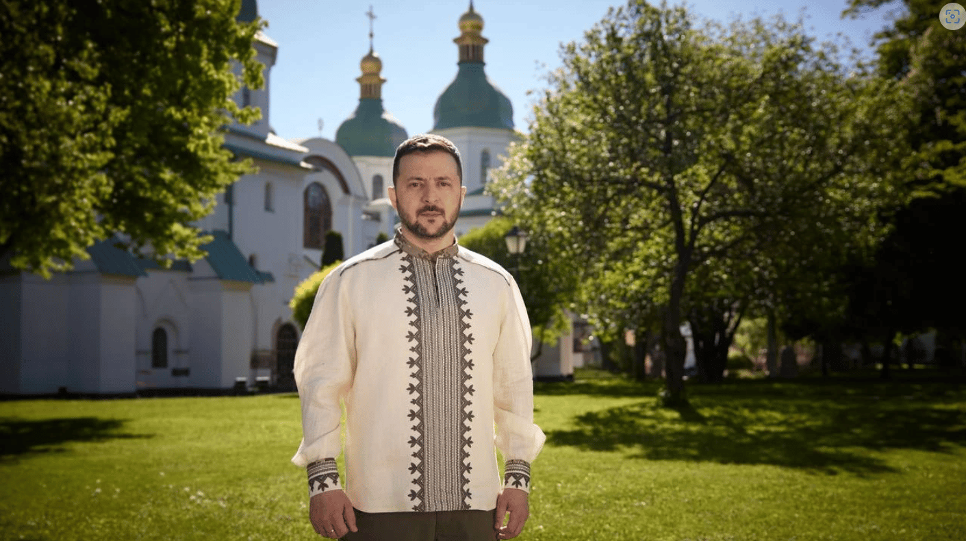 Mężczyzna z krótkimi ciemnymi włosami, w kremowej tradycyjnej koszuli z niebieskim haftem przez środek i czarnych spodniach stoi na trawniku obok zieleniącego się już wiosennie drzewa na tle cerkwii w Kijowie; cerkiew ma dwie wieże, których kopuły są szare i pokryte złotem.
