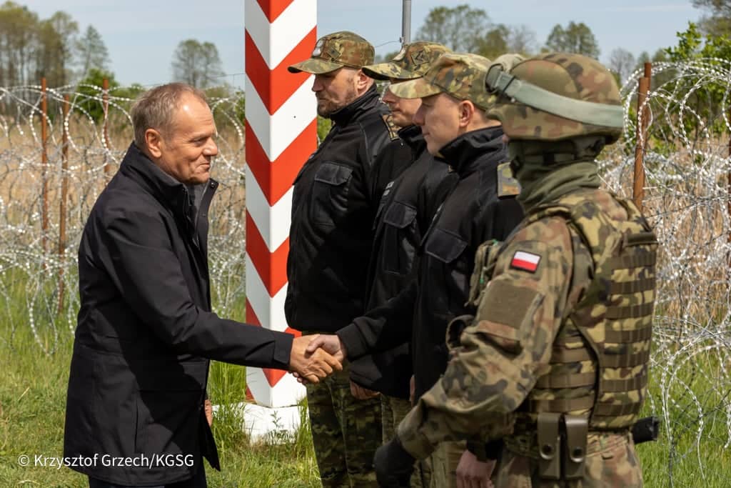mężczyzna w kurtce (Tusk) wita się z żołnierzami przy słupie granicznym