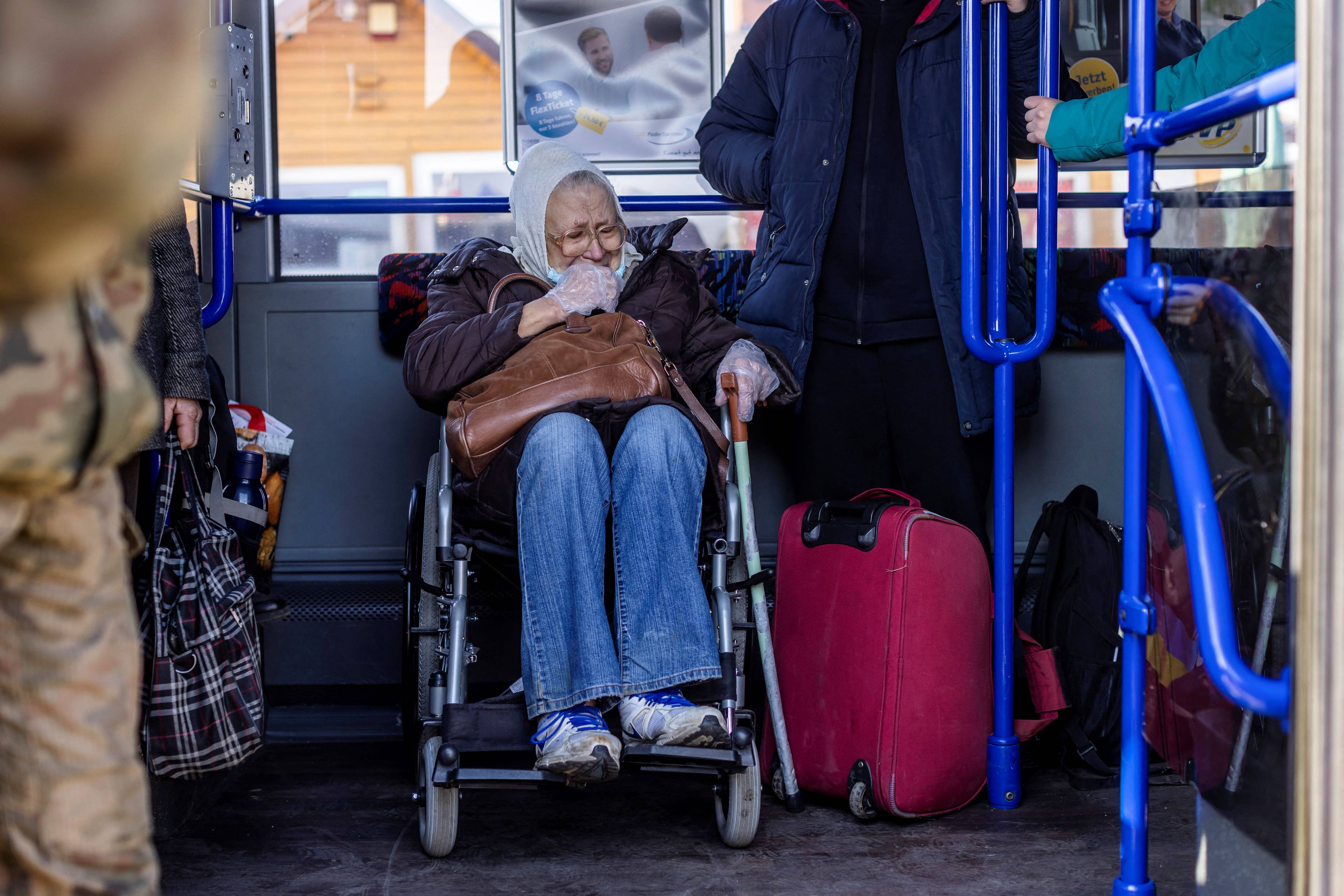 Na zdjęciu jest starsza pani z Ukrainy. Jest w wóxku inwalidzkim. Wygląda na przerażoną. Na głowie ma białą chustę, jest ubrana w ciemnobrązową kurtkę i dżinsy