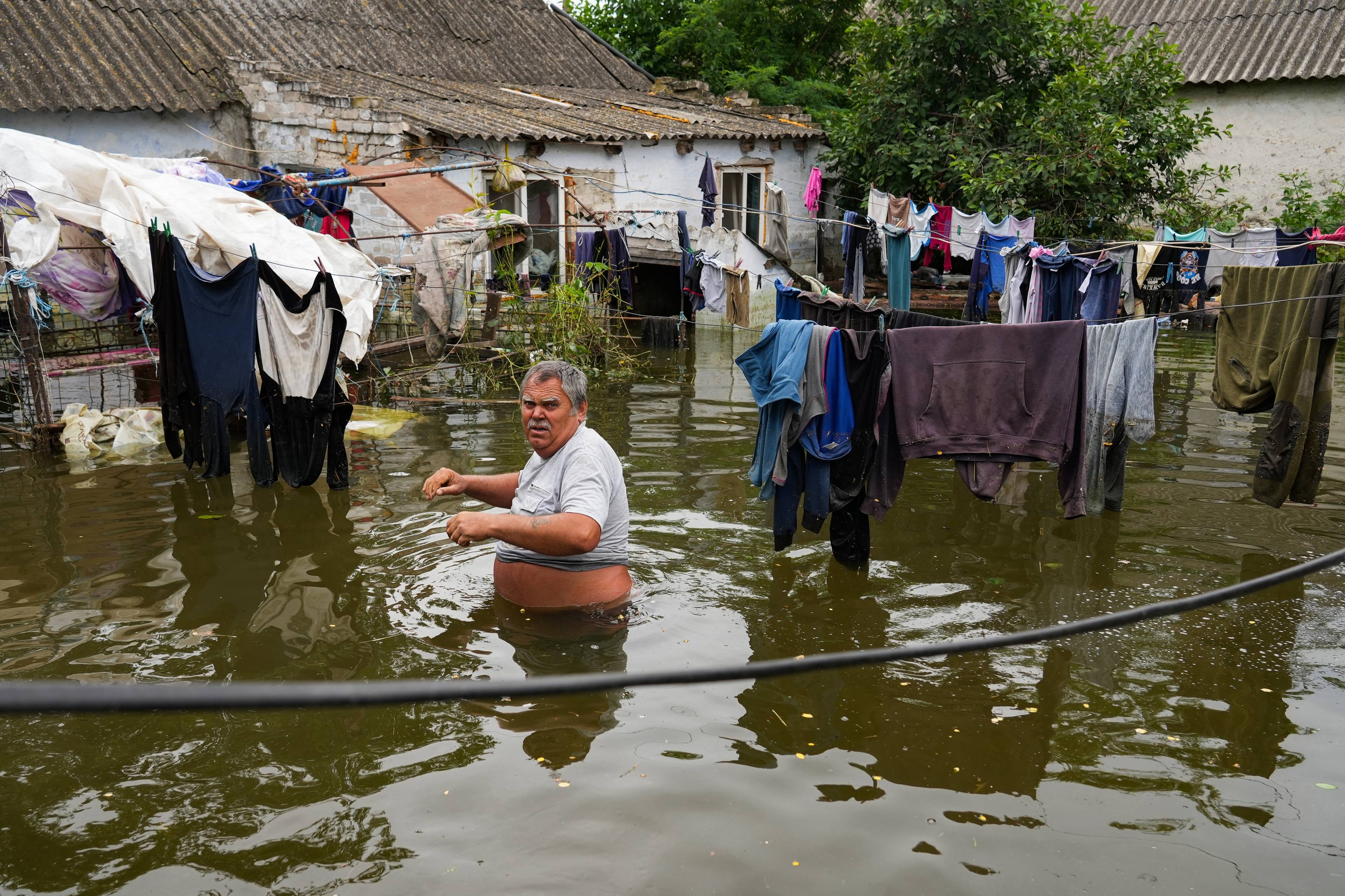 Na zdjęciu jest starszy mężczyna. Do pasa jest stoi w wodzie. Z tyłu widać jego zatopiony dom, obok schną rozwieszone na linach rzeczy