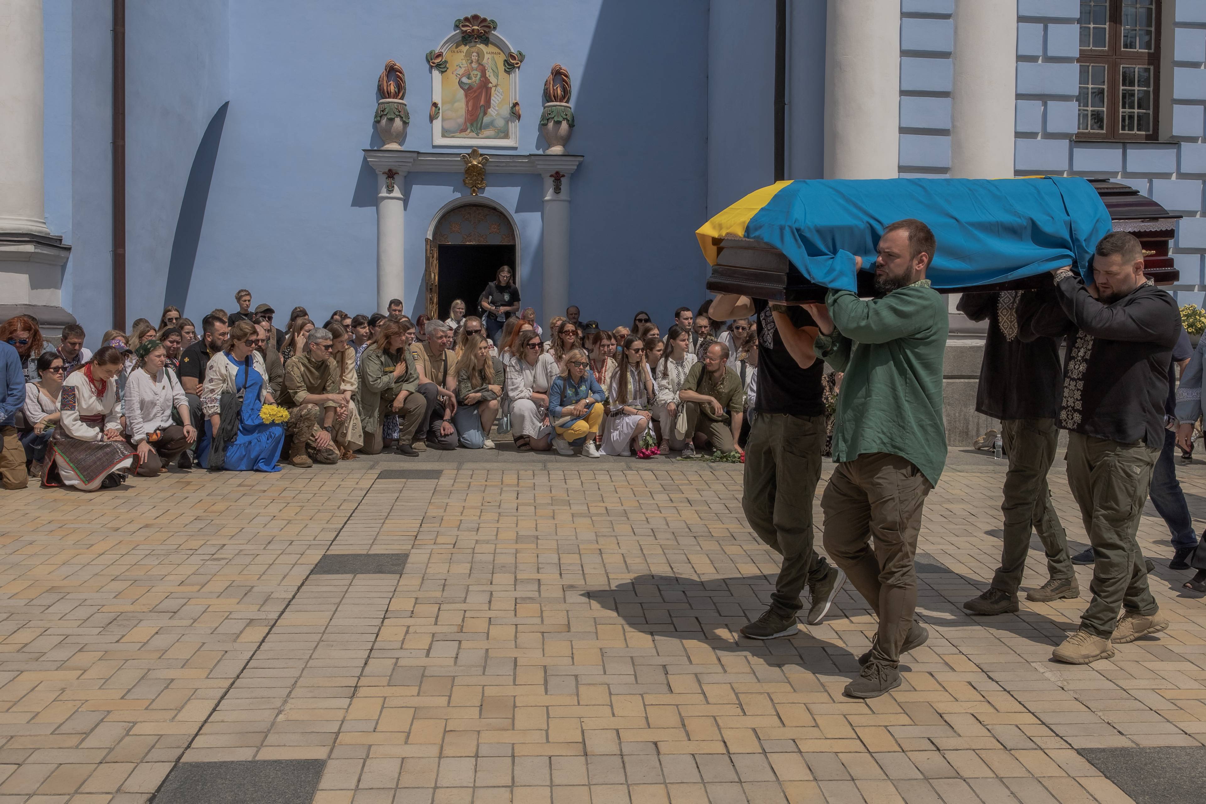 mężczyźni niosą trumnę spowitą w niebiesko-zółtą flagę, w tle cerkiew i klęczący żałobnicy