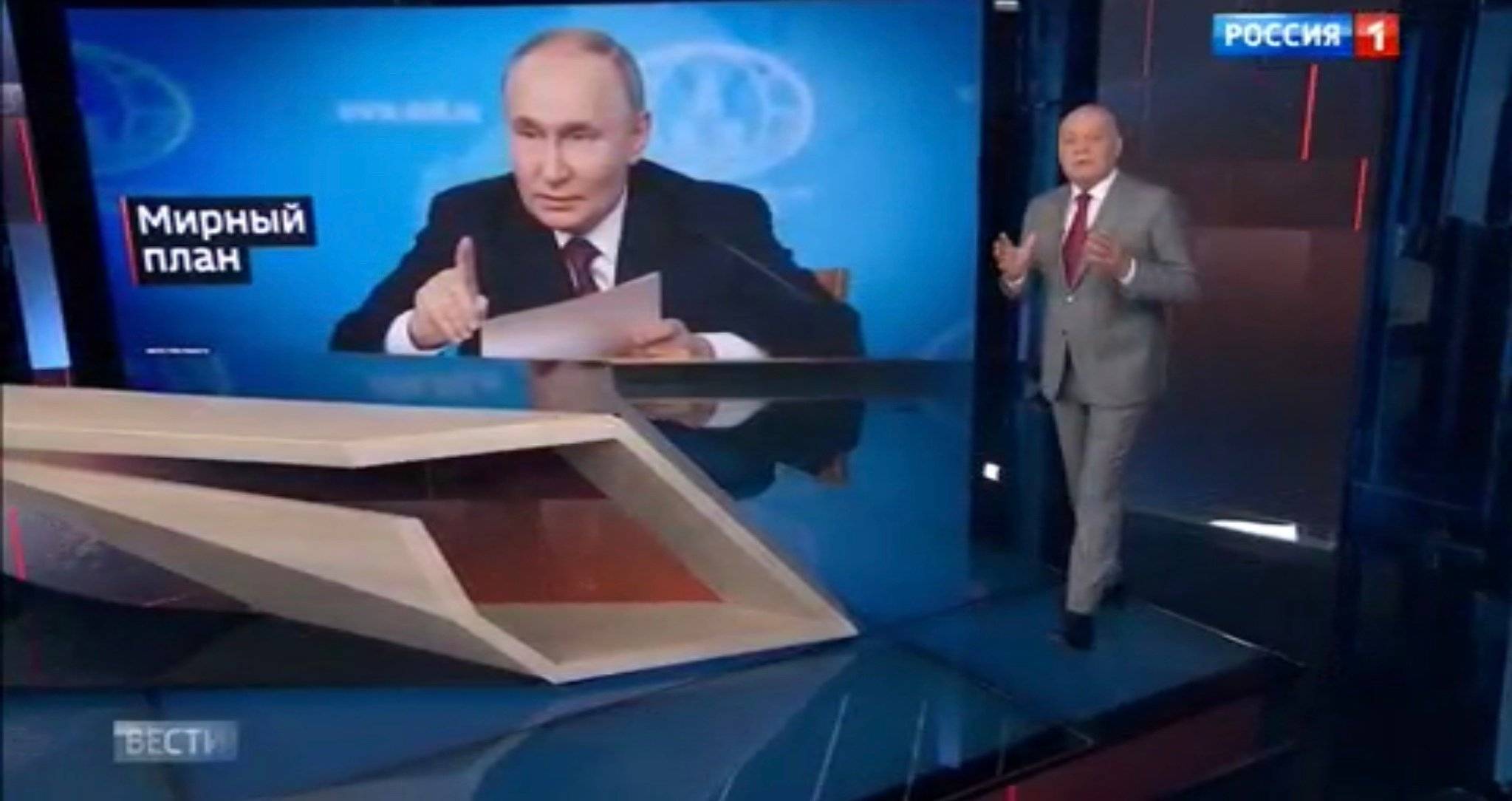 Studio telewizyjne. Prezenter - starszy pan (Dmitrij Kisielow) gestykuluje. na ekranie - zdjęcie {utina czytającego z kartki
