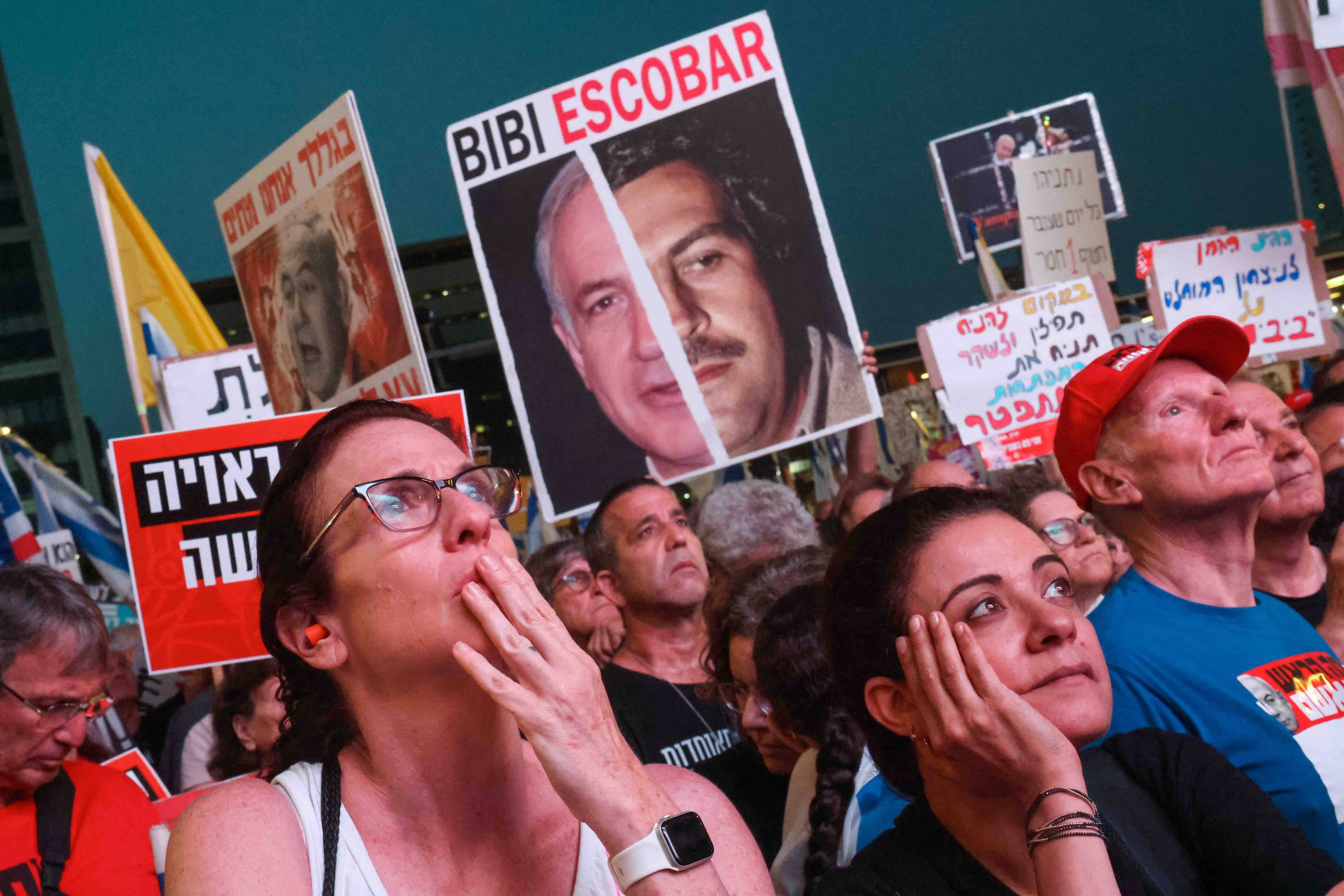 Protestujący na ulicy Tel Awiwu ludzie, w tle plakat przedstawiający podobiznę premiera Izraela Binjamina Netanjahu, połowa twarzy przysłonięta jest zdjęciem Pablo Escobara, przywódcy kartelu narkotykowego,