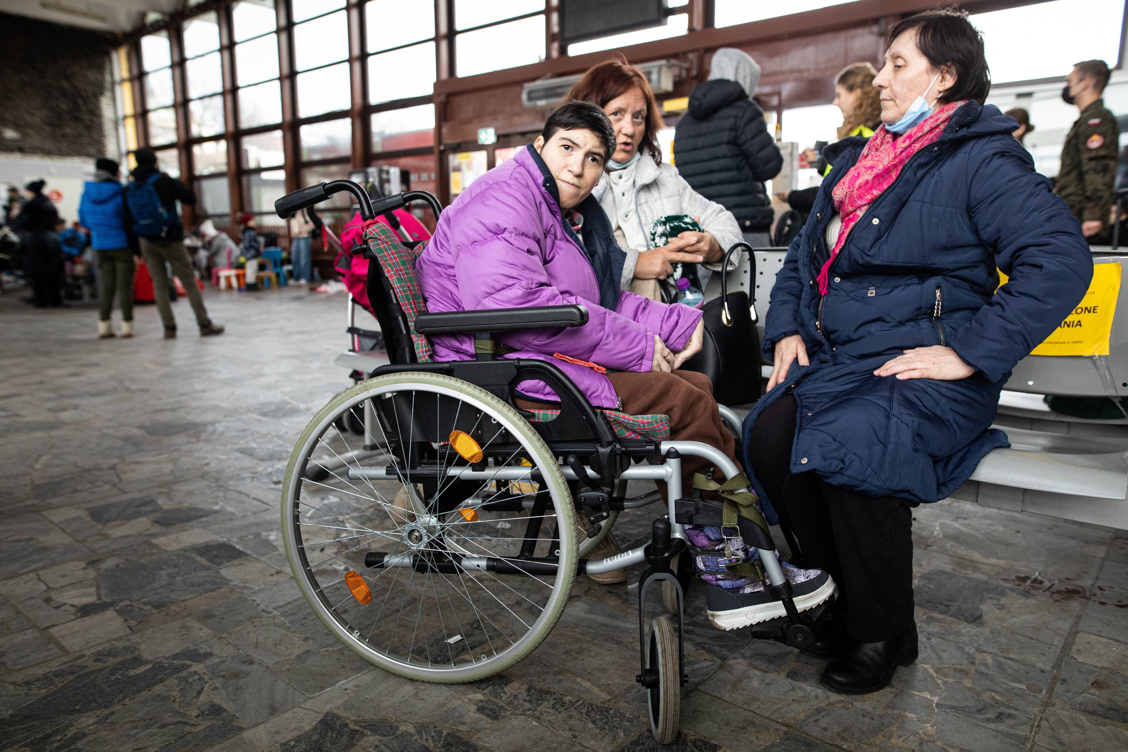 Na zdjęciu jest trzy kobiety. Jedna z nich siedzi w wózku inwalidzkim. Jest ubrana w kurtkę fioletową.