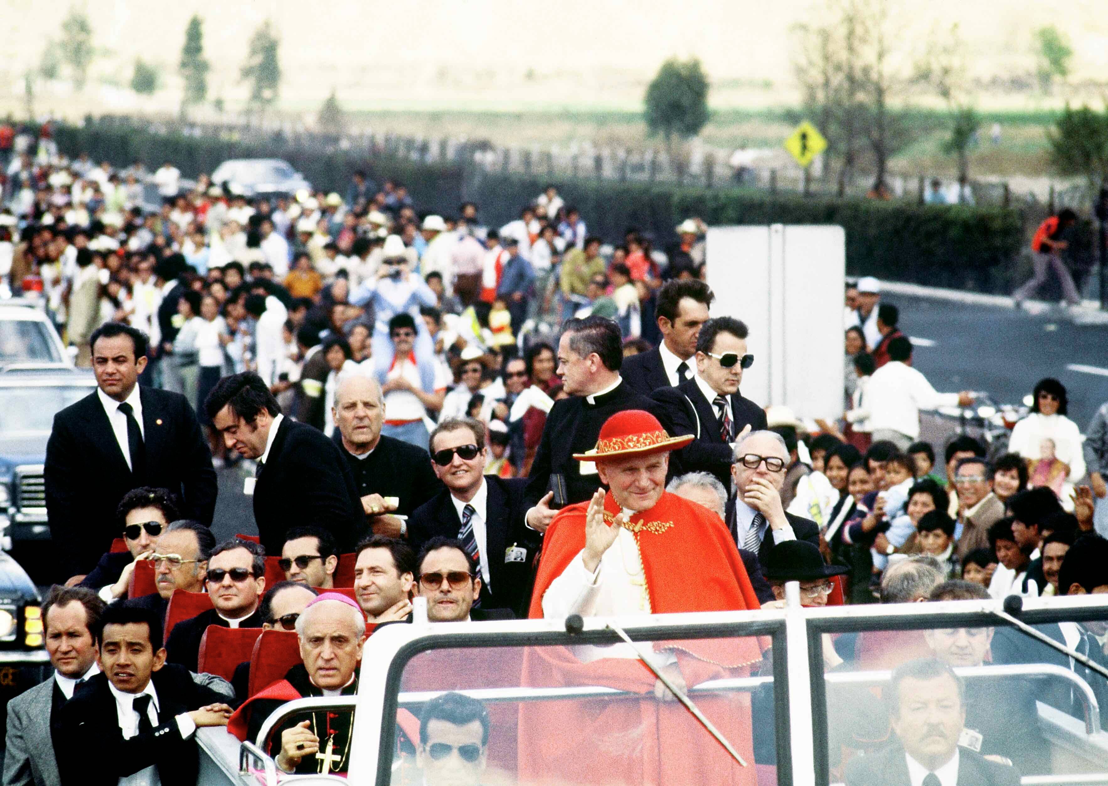Papież Jan Paweł II w czerwonym kapeluszu jedzie samochodem ulicą, otoczony przez tłum,. w drugim rzędzie za papieżem siedzi biskup Marcinkus. Watykan