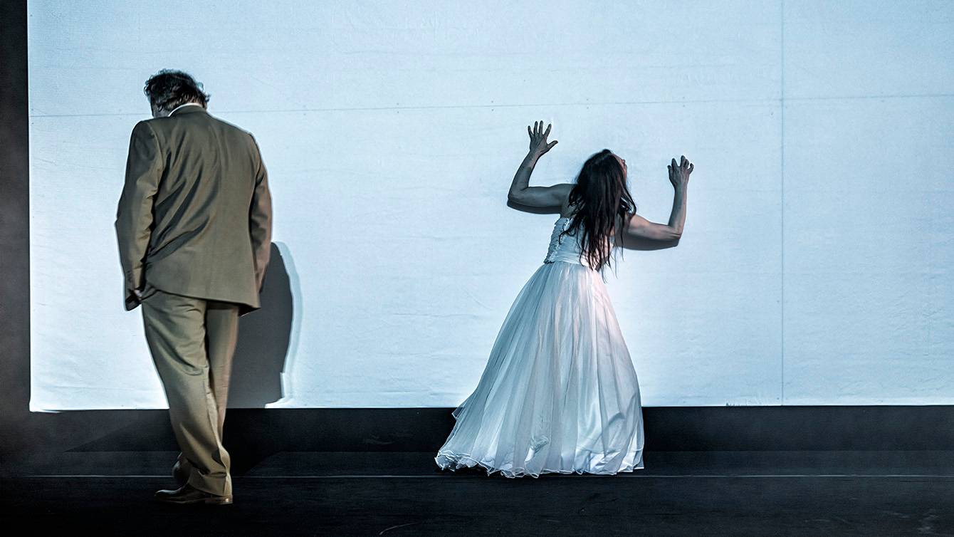 Scena ze spektaklu „Pewnego długiego dnia”, sylwetki stojących tyłem do obiektywu mężczyzny w marynarce i kobiety w białej sukni na tle białego ekranu