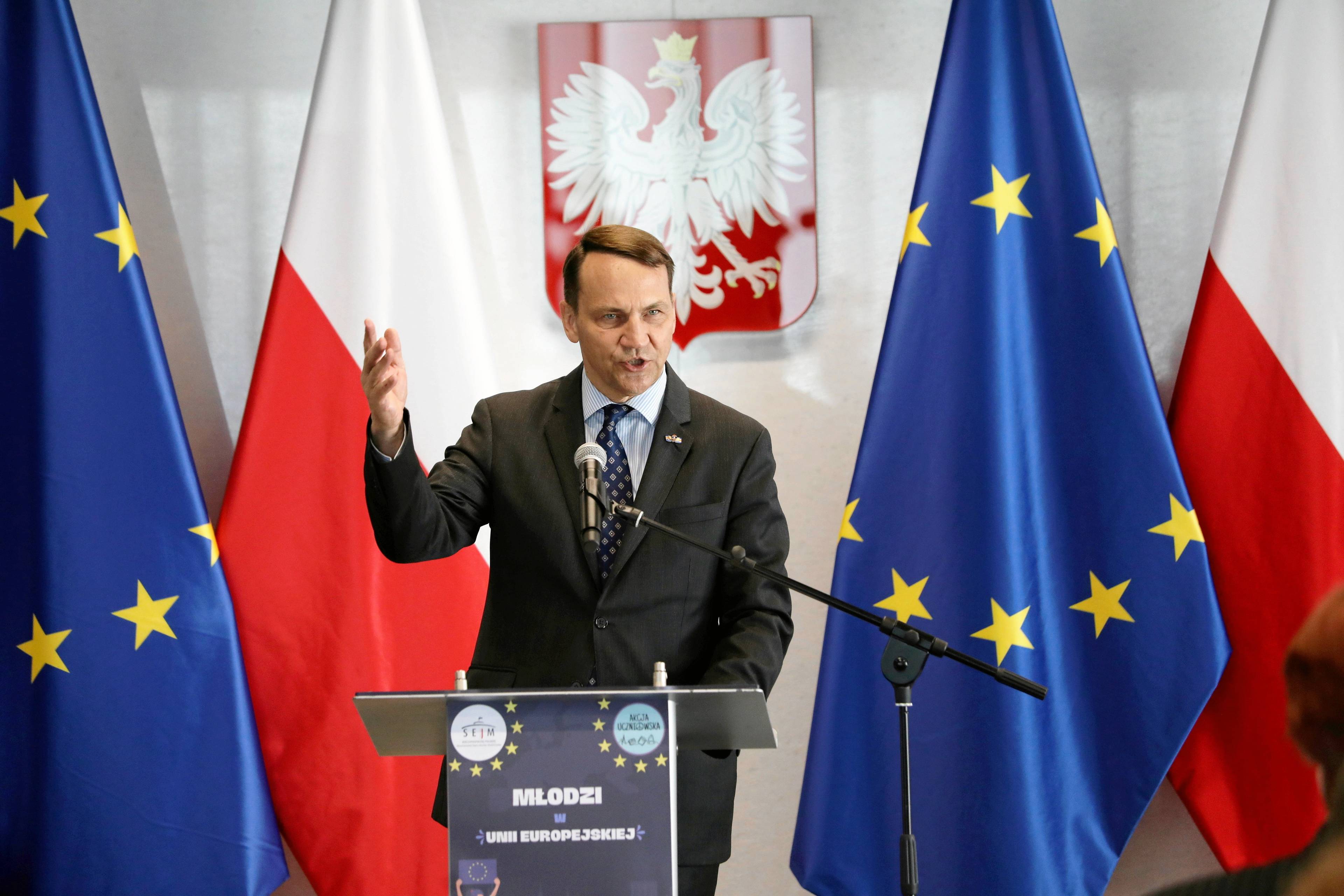 Polityk w garniturze z podniesioną ręką przemawia na tle flag Polski i Unii