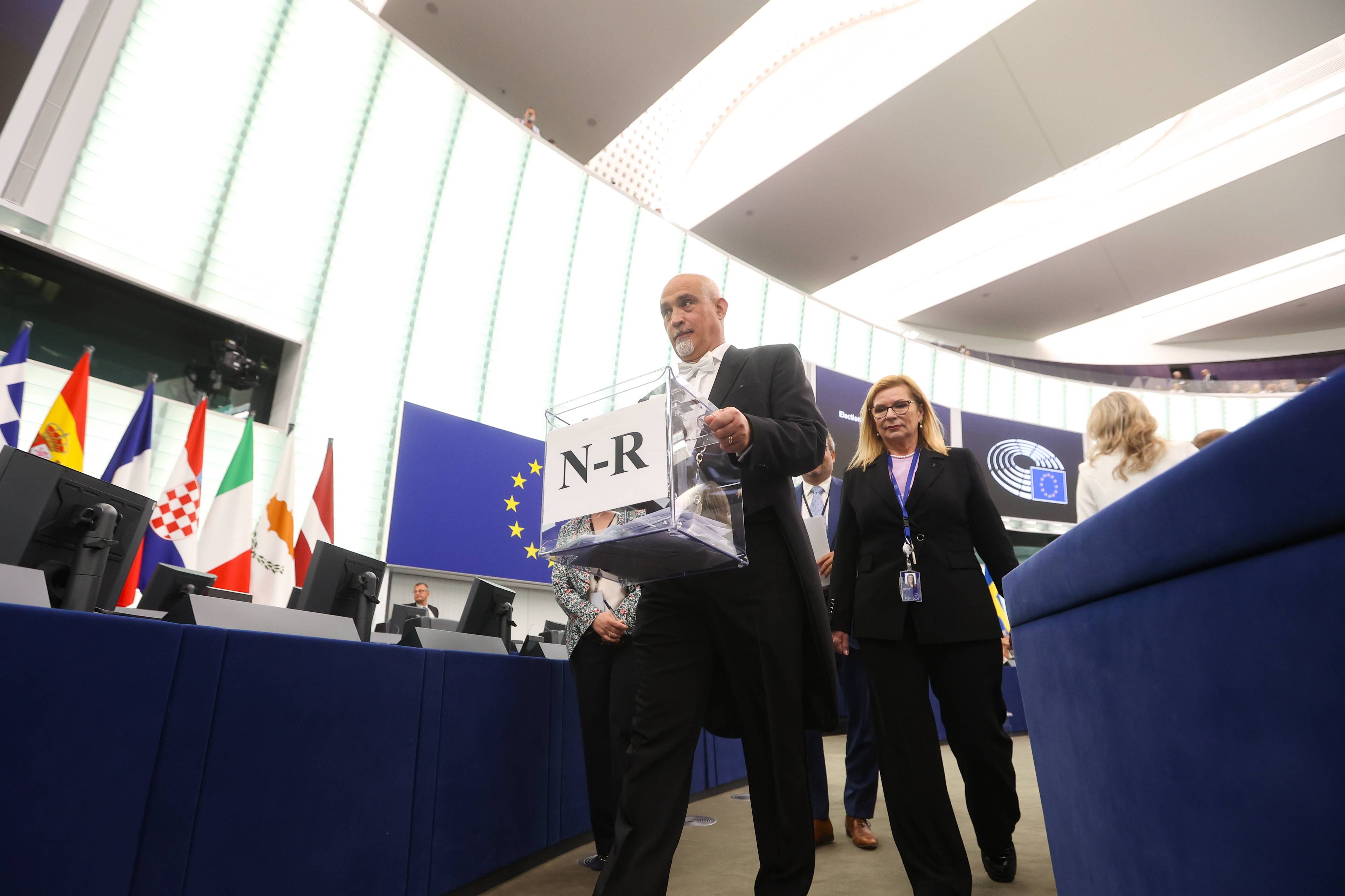 Mężczyzna w czarnym fraku niesie przezroczystą urnę wyborczą, w tle flaga europejska i flagi narodowe państw członkowskich UE