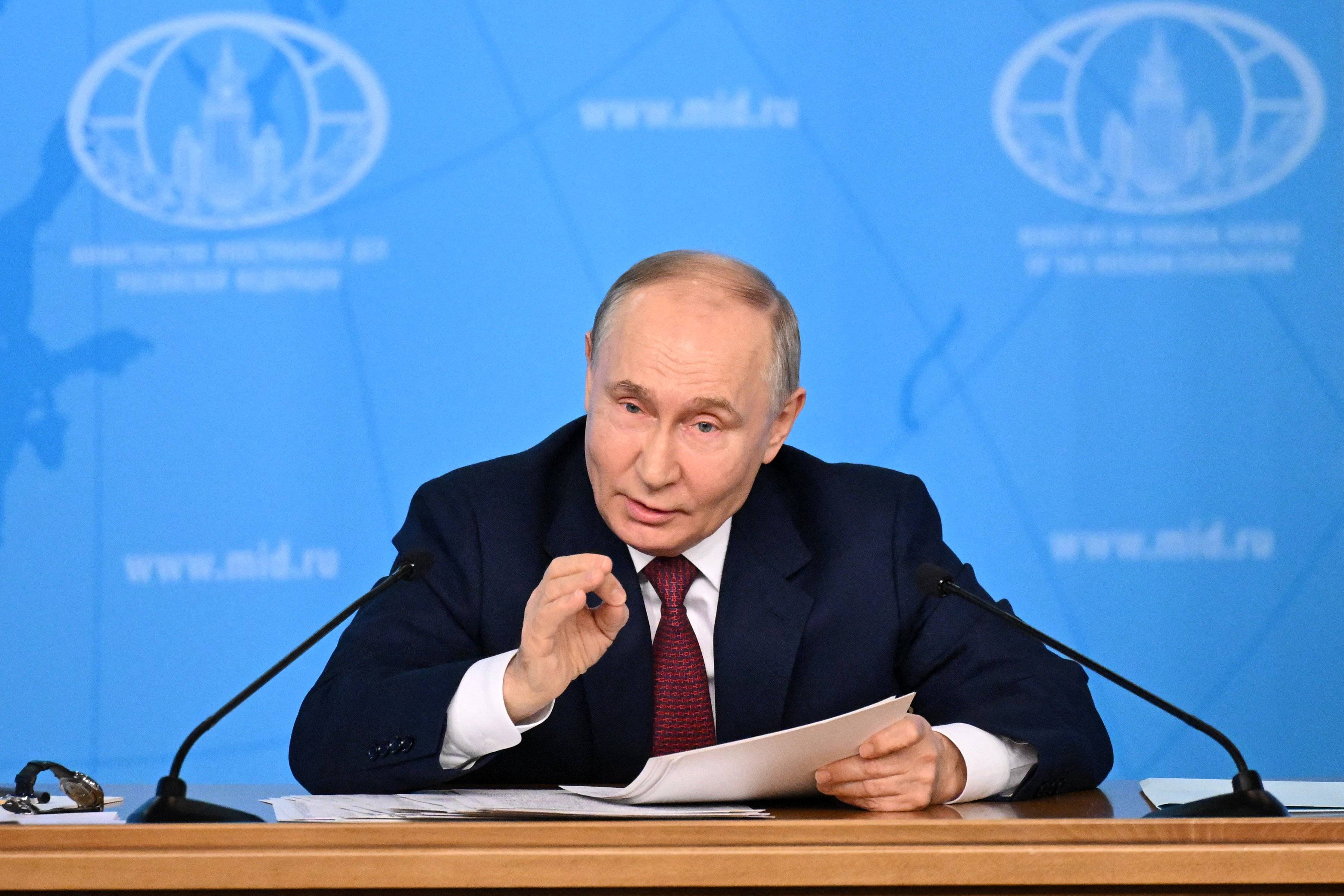 Władimir Putin. Łysiejący, krępy mężczyzna w garniturze i białej koszuli, siedzi przy stole, gestykuluje jedną dłonią.