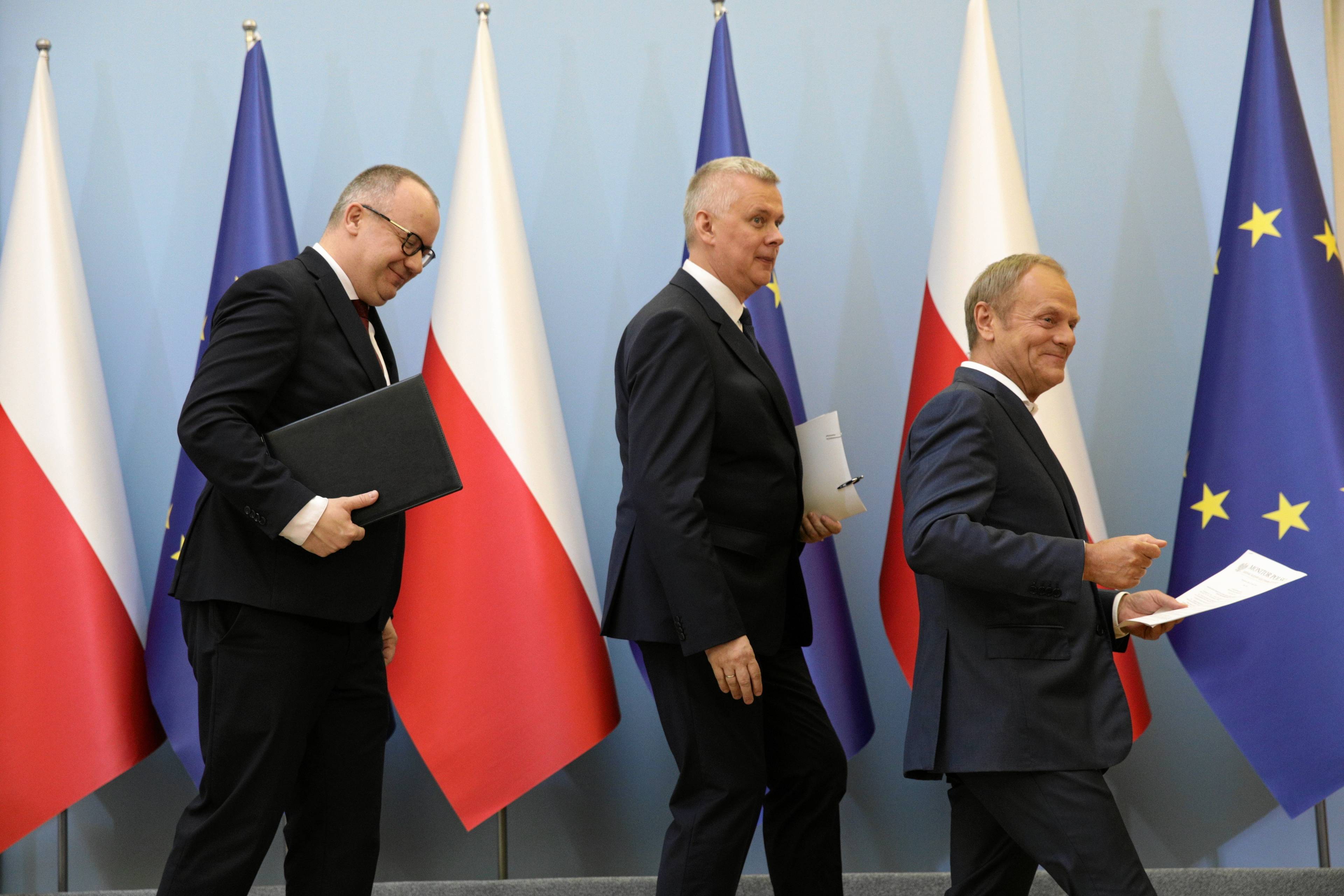 Trzech mężczyzn w garniturach na tle polskich i unijnych flag: Adam Bodnar, Tomasz Siemoniak i Donald Tusk