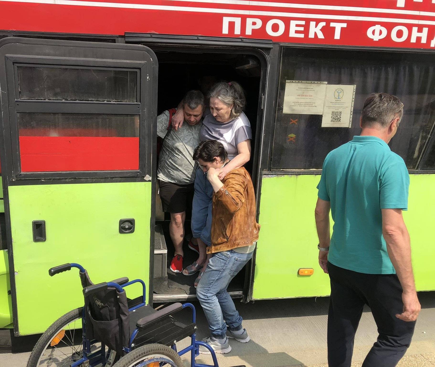 Na zdjęciu dwóch mężczyzn pomaga wyjść starszej pani z zielonego autobusu. Obok stoi wózek inwalidzki