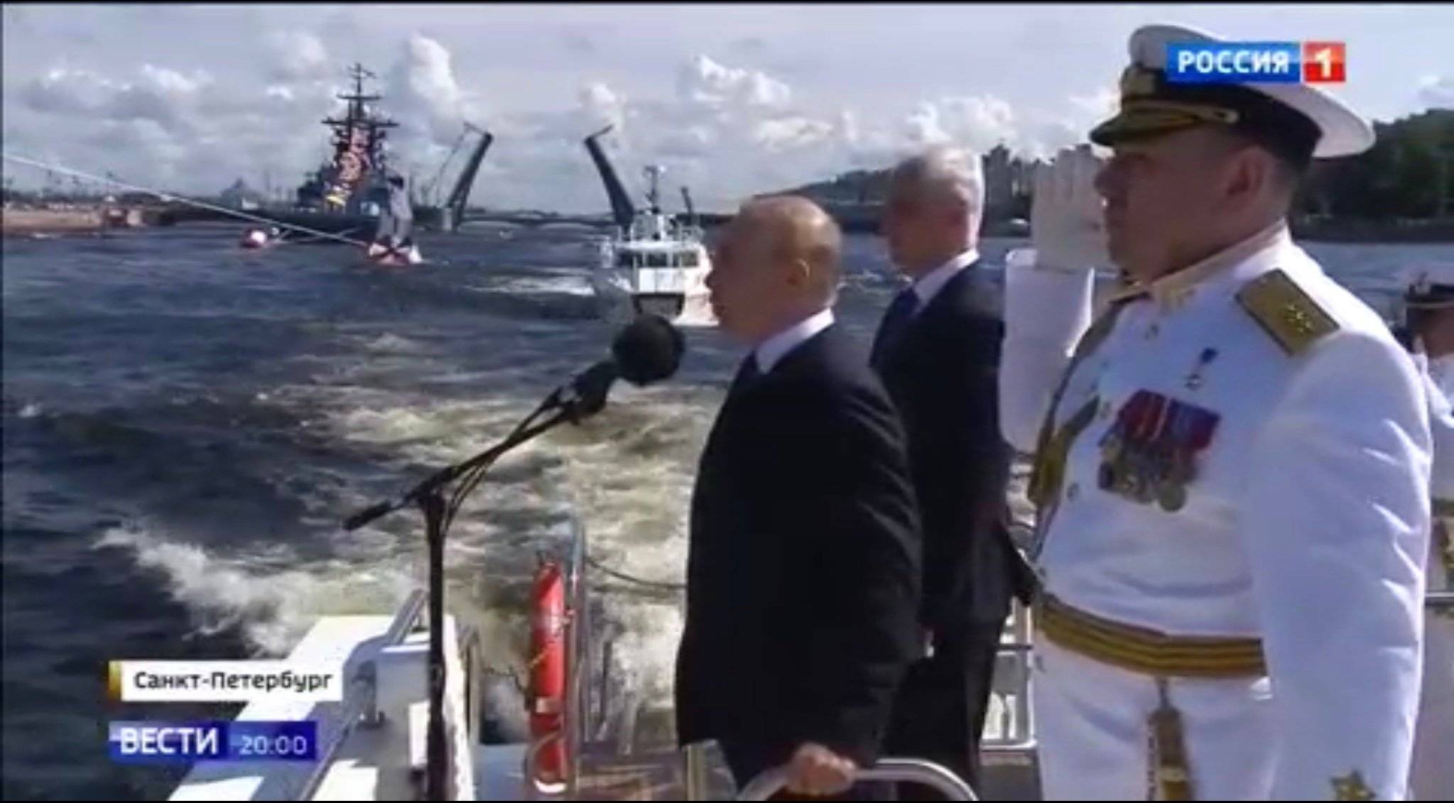 Putin mówi do mikrofonu stojąc w kutrze w towarzystwie oficerów floty w białych mundurach