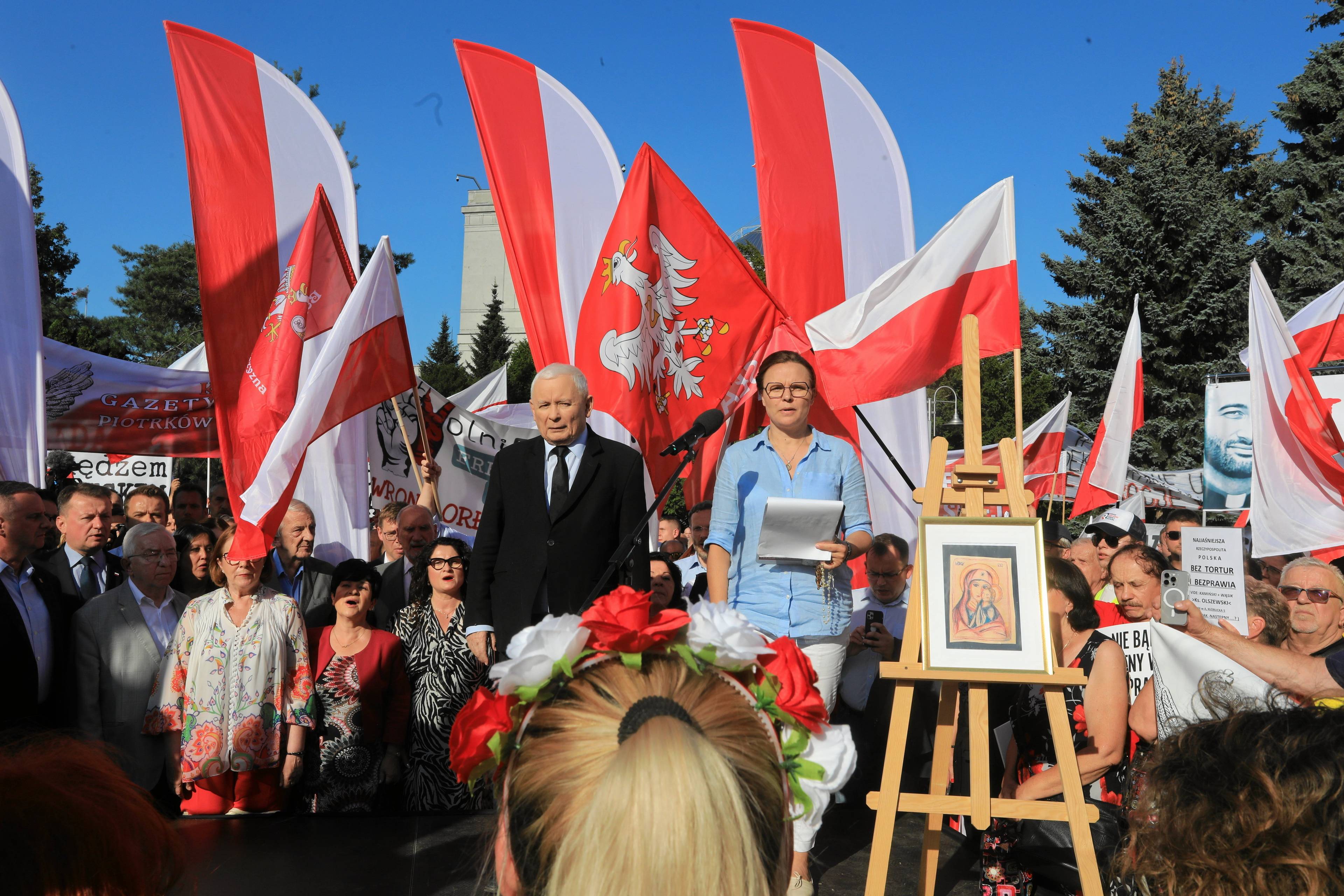 Jarosław Kaczyński stoi na scenie w pełnym słońcu, ob niego kobieta, za nimi polskie flagi