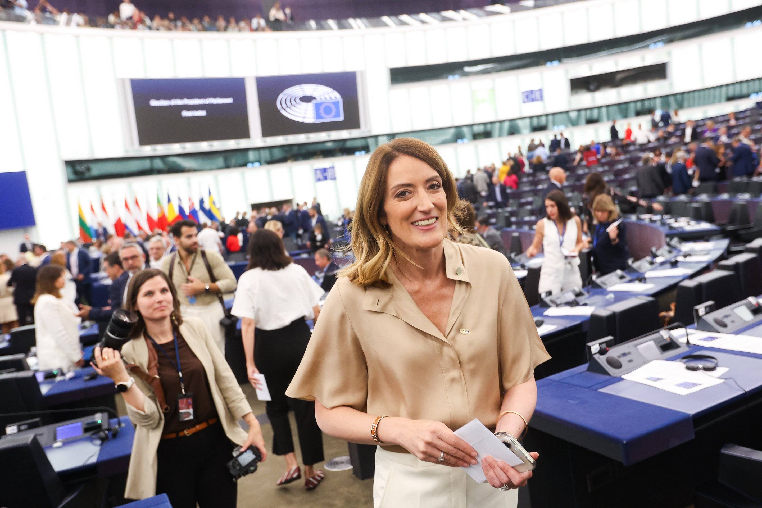 Uśmiechnięta kobieta z półdługimi włosami w kremowej bluzce ze złożoną kartką papieru w dłoniach idzie w kierunku urny wyborczej w dużej i przestronnej sali plenarnej Parlamentu Europejskiego, wokół niej widać wiele osób