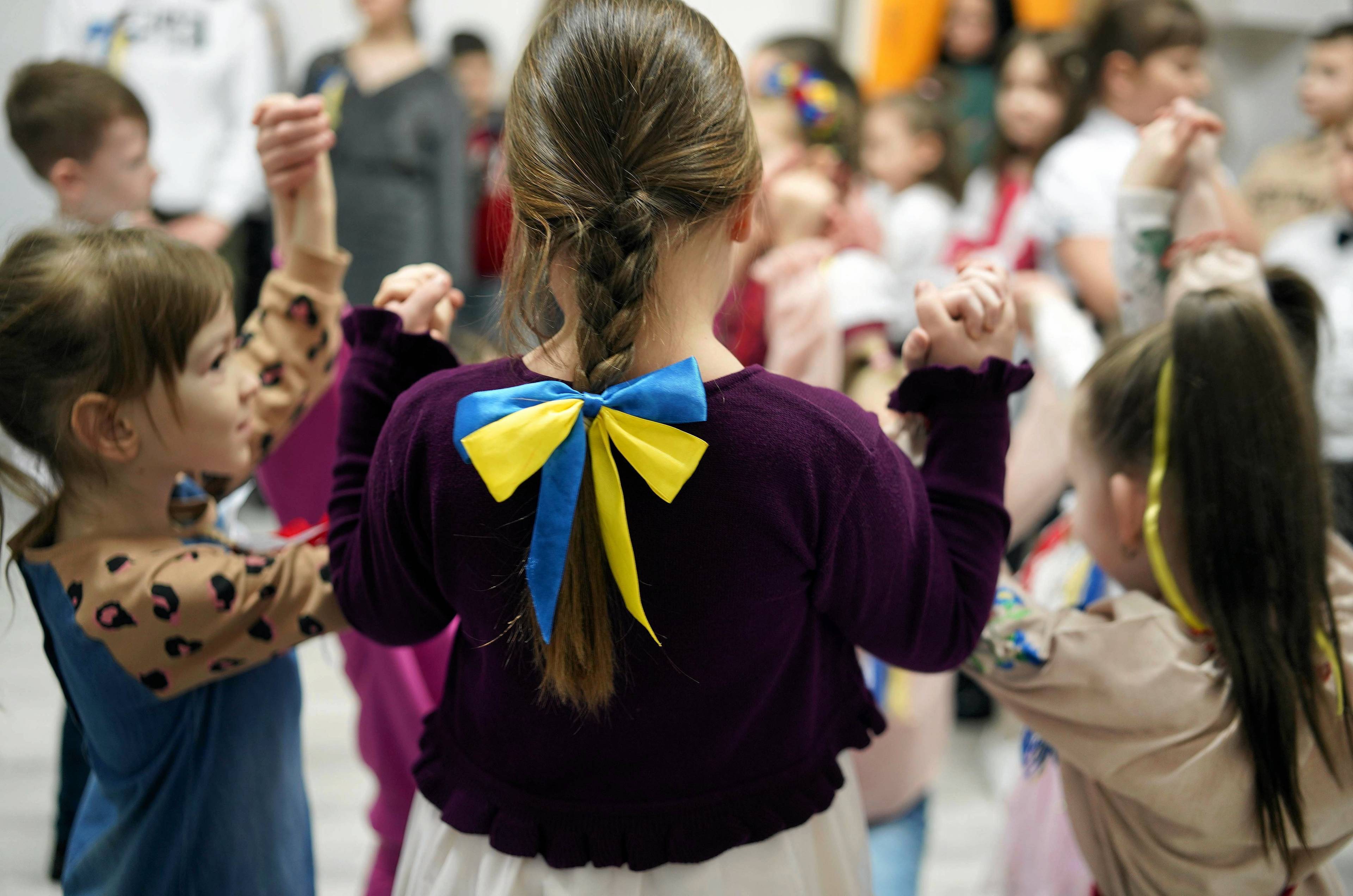 Na zdjęciu są dzieci w wieku 6-7 lat. Po środku tyłem stoi dziewczynka. Jest ubrana w bordową bluzkę, na wakoczyku ma zawiązaną kokardę w kolorach niebiesko-żółtych. Ma brązowe włosy