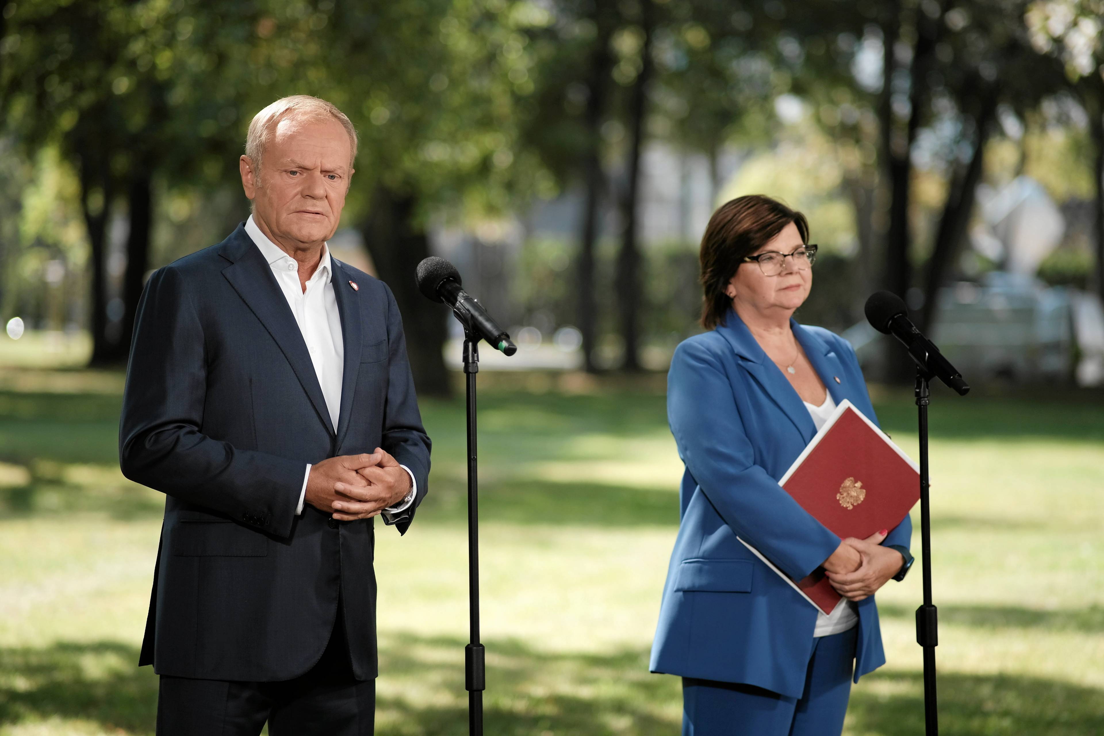 Donald Tusk i ministra zdrowia Izabela Leszczyna stoją przed mikrofonami w parku podczas konferencji prasowej. Wytyczne w sprawie aborcji