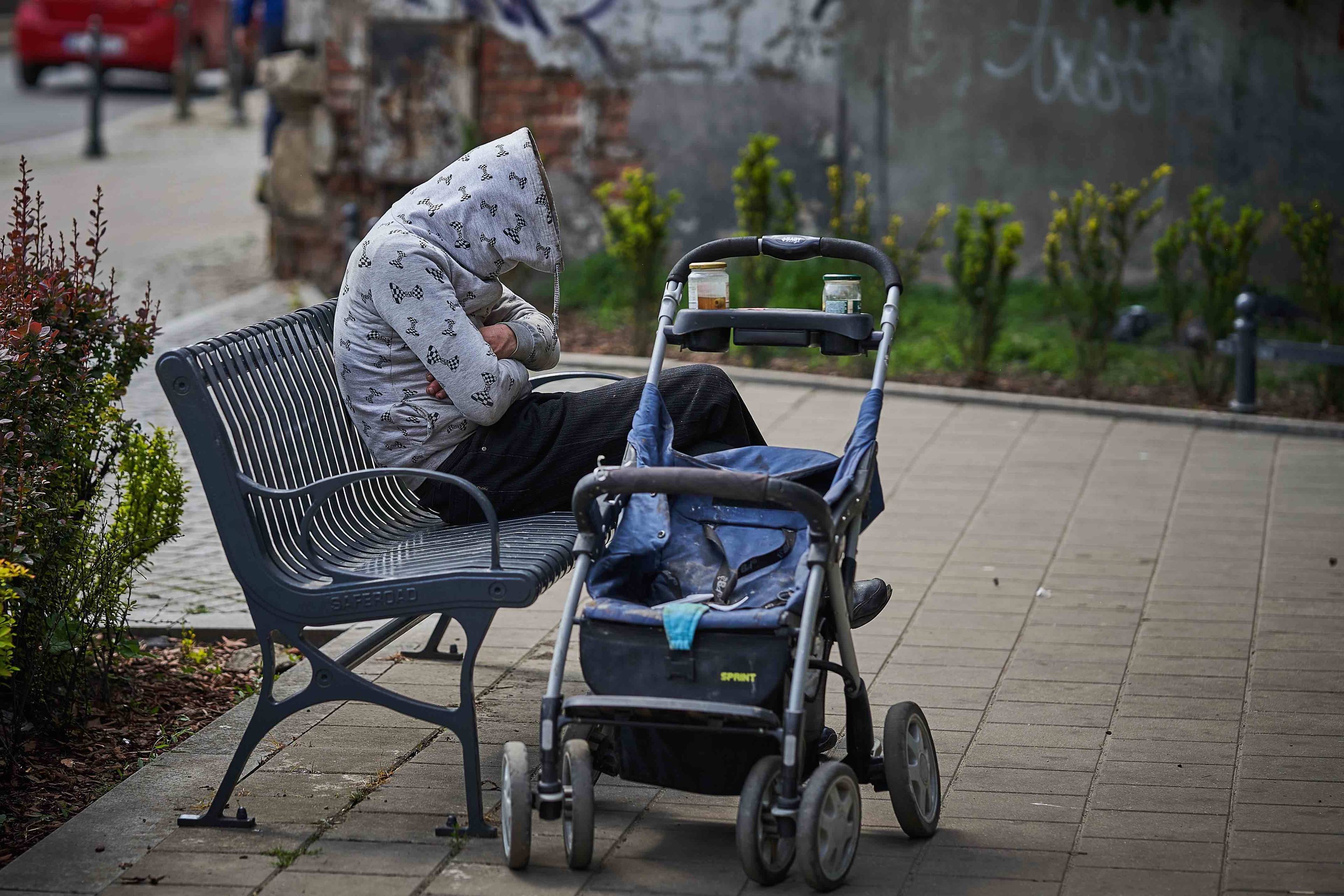 Osoba bezdomna siedzi na ławce, przed nią stoi wózek dziecięcy.