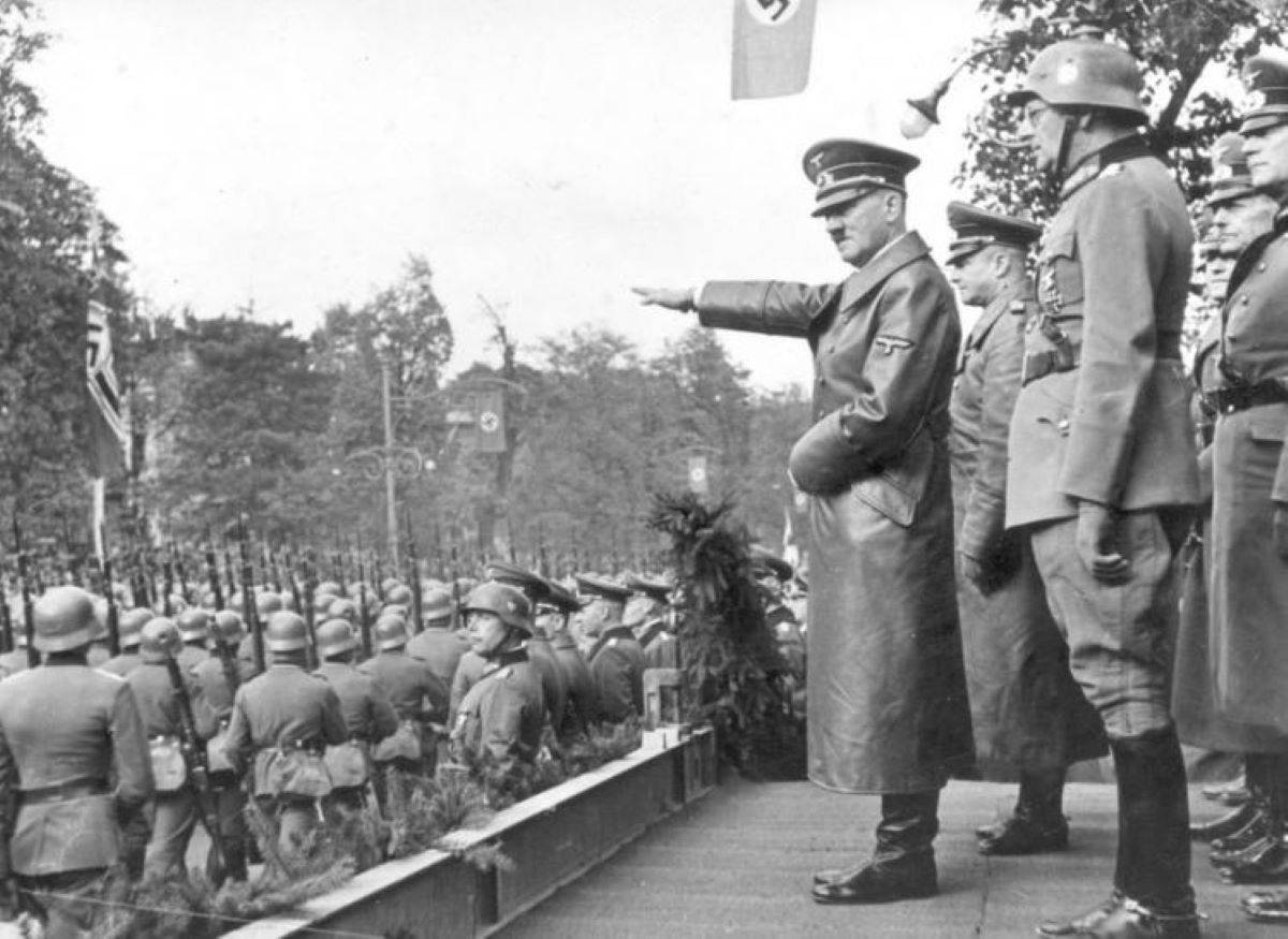 Archiwalne czarno-białe zdjęcie, na którym Adolf Hitler w wojskowym uniformie salutuję podniesioną ręką wojsko maszerujące ulicą