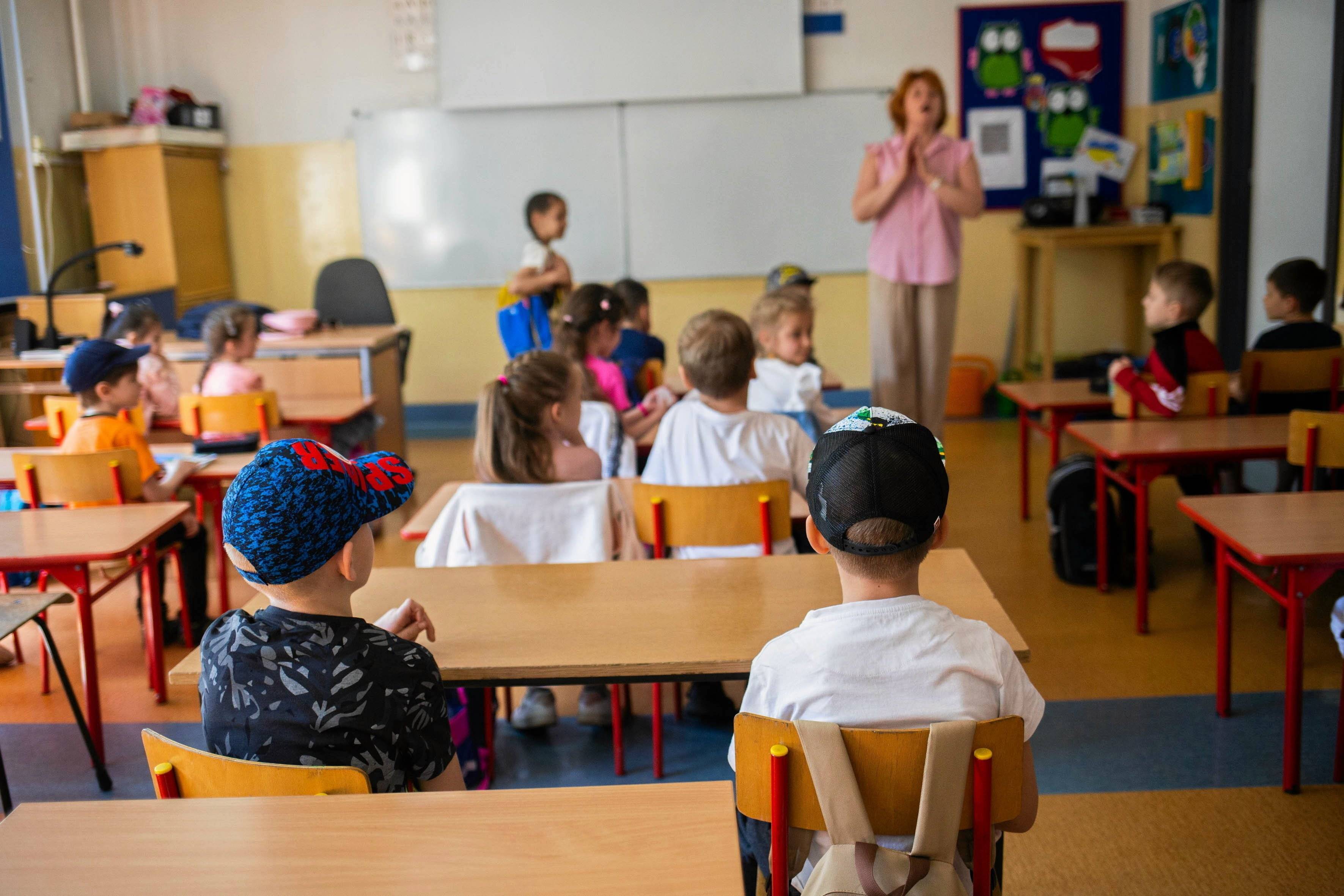 Na zdjęciu są dzieci w wieku ok. siedem lat. Siedzą za szkolnymi ławkami. W tle obok tablicy jest nauczycielka. Ma różową bluzkę i rude włosy