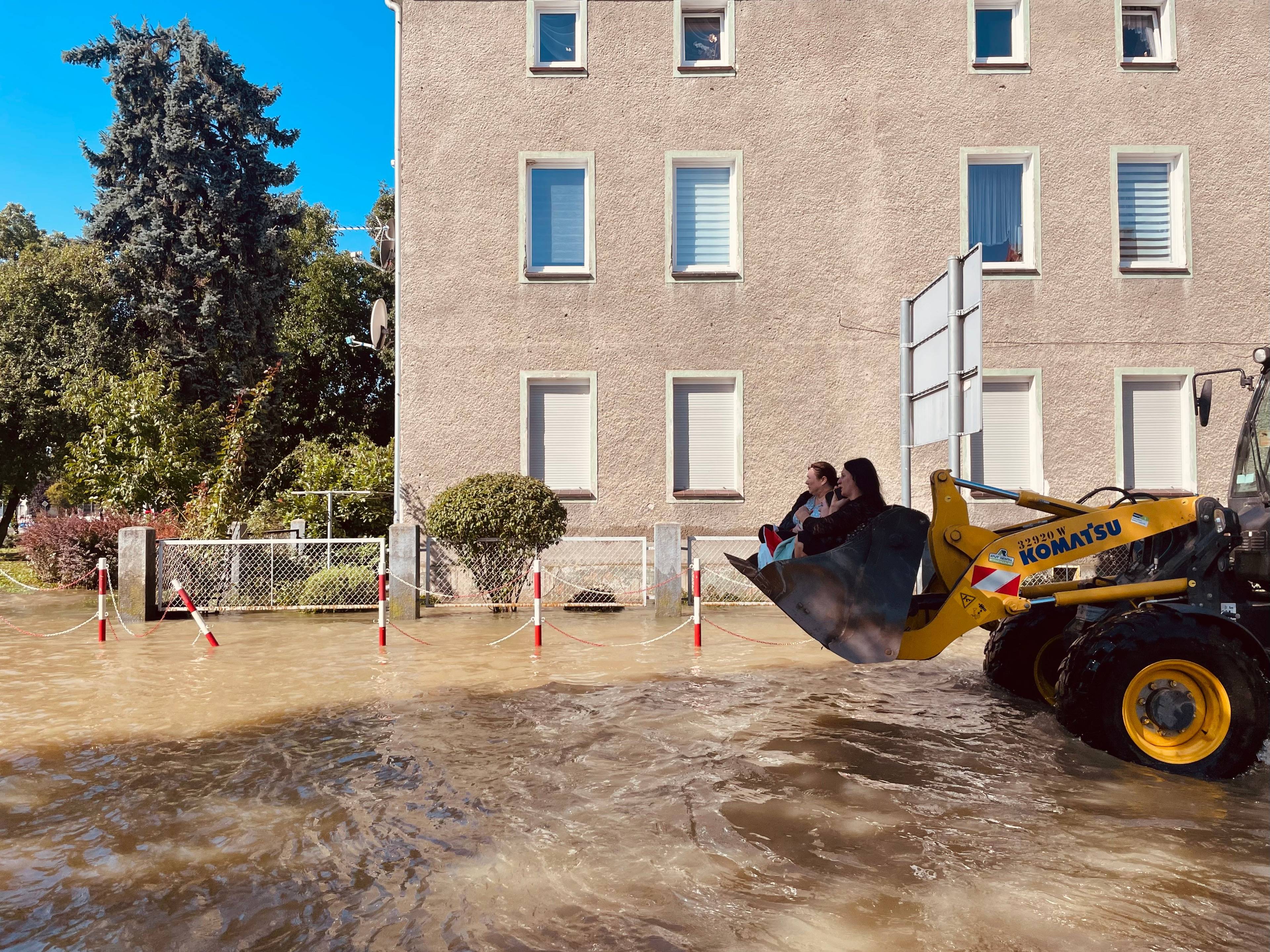 Kobiety siedzą w łyżce koparki, która jedzie zalaną wodą ulicą