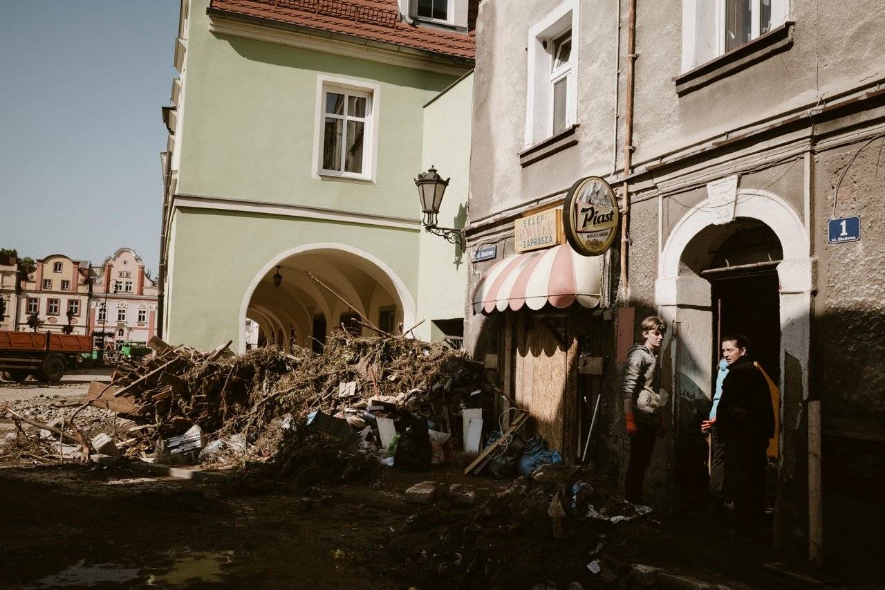 Góra pni i kłód tarasuje wejście do podcieni na zabytkowym rynku, w bramie stoi chłopak i kobieta