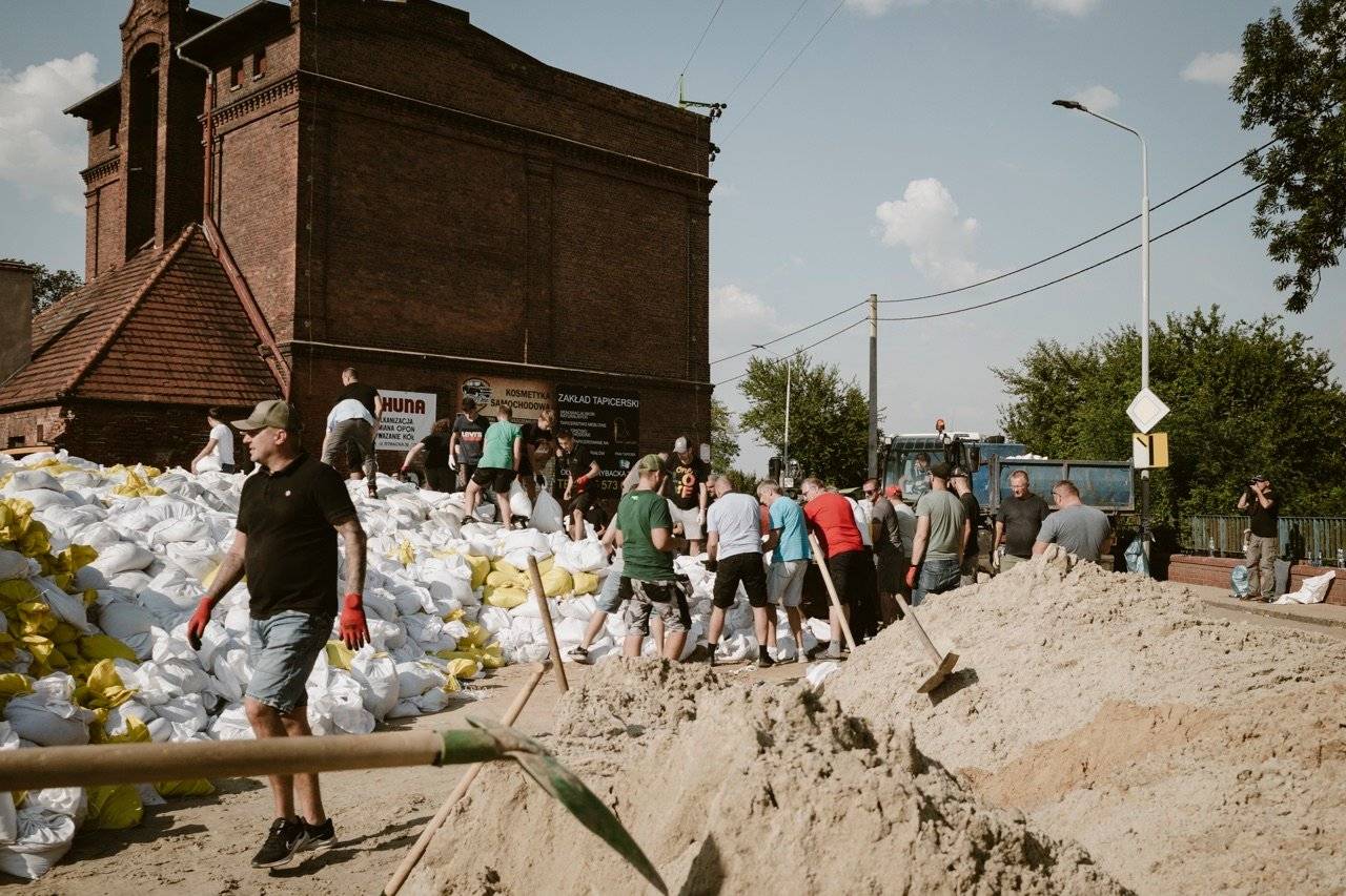 Ludzie budują wał z worków z piaskiem, w tle kamienica, na pierwszym planie pryzma piasku i łopata