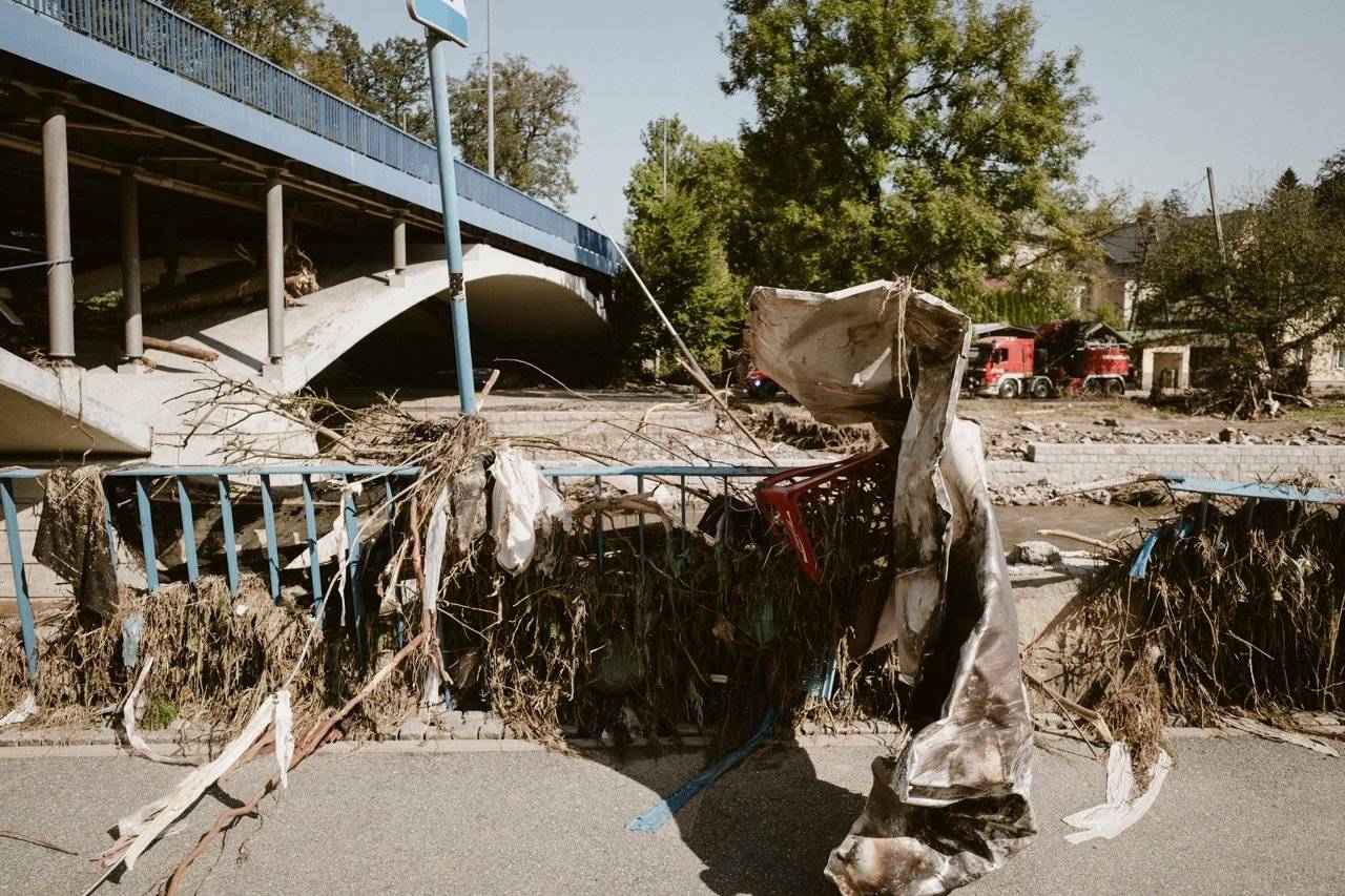 Most przez rzekę, na pierwszym planie konary drzew i pnie na metalowej barierce, na drugim brzegu rzeki stoi wóz strażacki